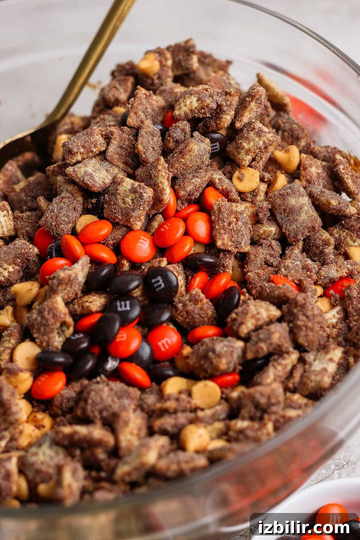 No-Bake Pumpkin Spice Protein Puppy Chow Recipe 4 A glass bowl filled with chocolate-coated cereal pieces, peanut butter chips, and red, orange, and brown candy-coated chocolates.