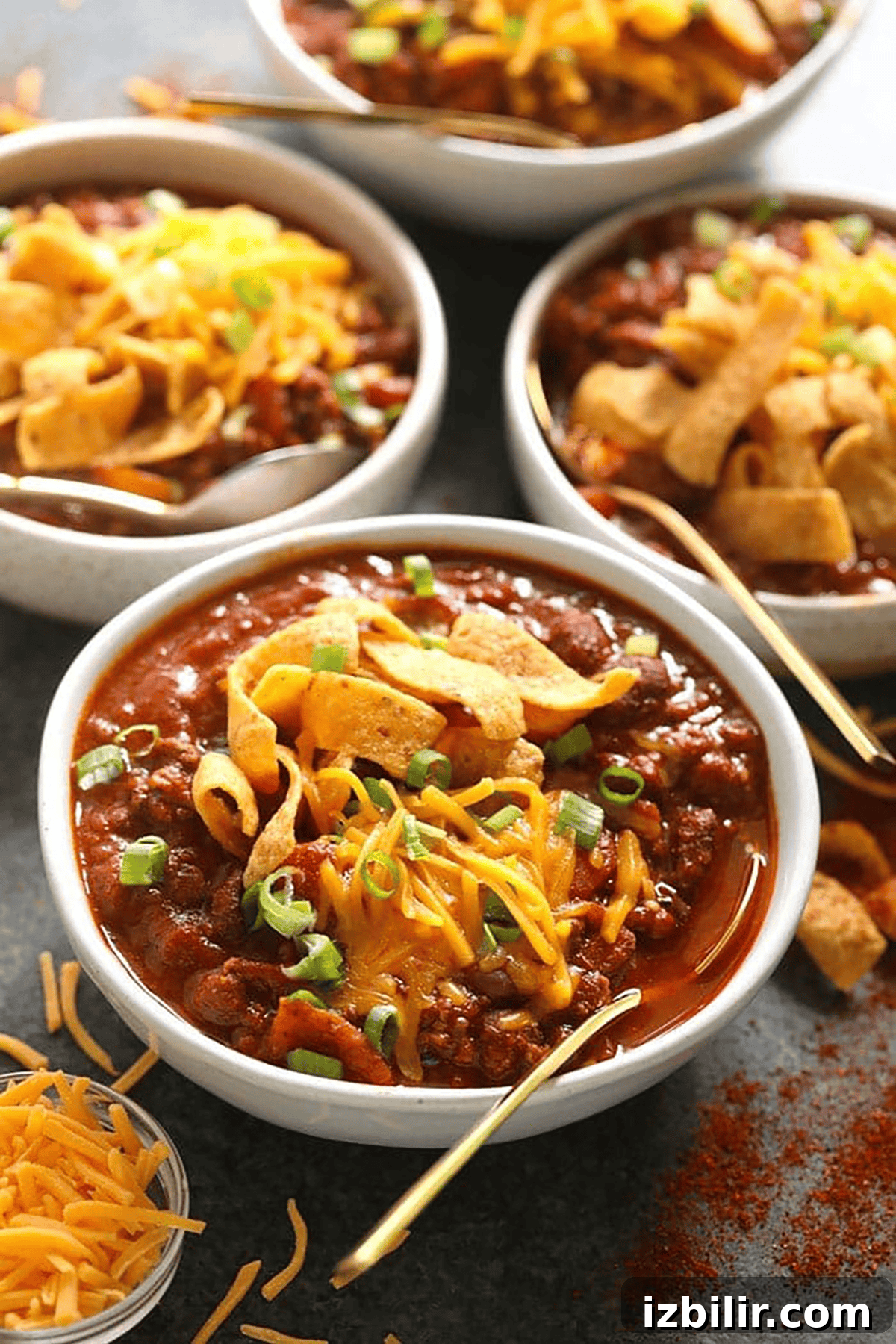 Award-Winning Chili Recipe That Won Multiple Cook-Offs 2 Four bowls of chili topped with shredded cheddar cheese, corn chips, and chopped green onions, with a side of shredded cheese and gold spoons.