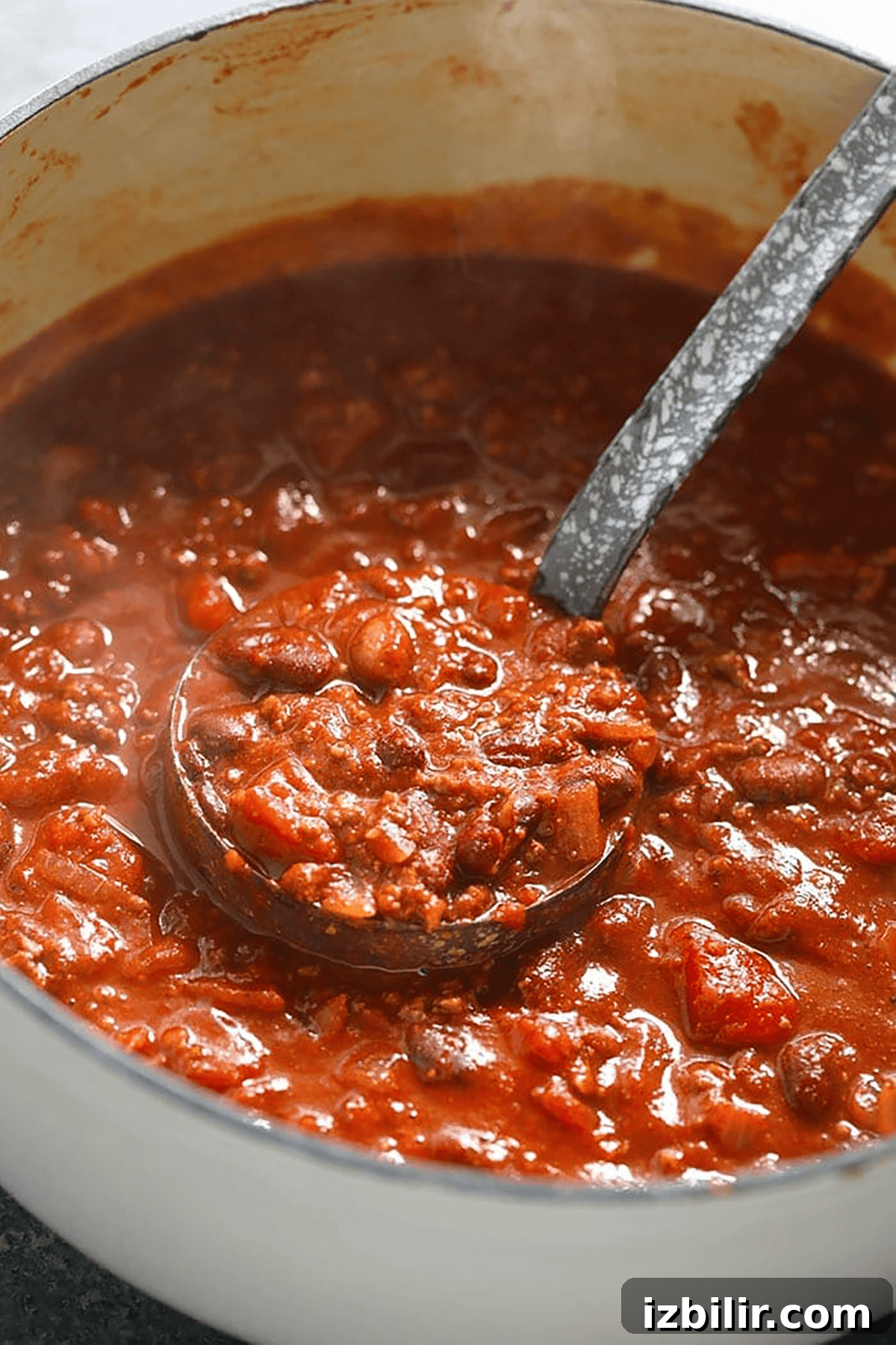 Award-Winning Chili Recipe That Won Multiple Cook-Offs 3 A ladle scoops thick chili with beans, tomato chunks, and ground meat from a large white pot—an inviting chili recipe in progress.