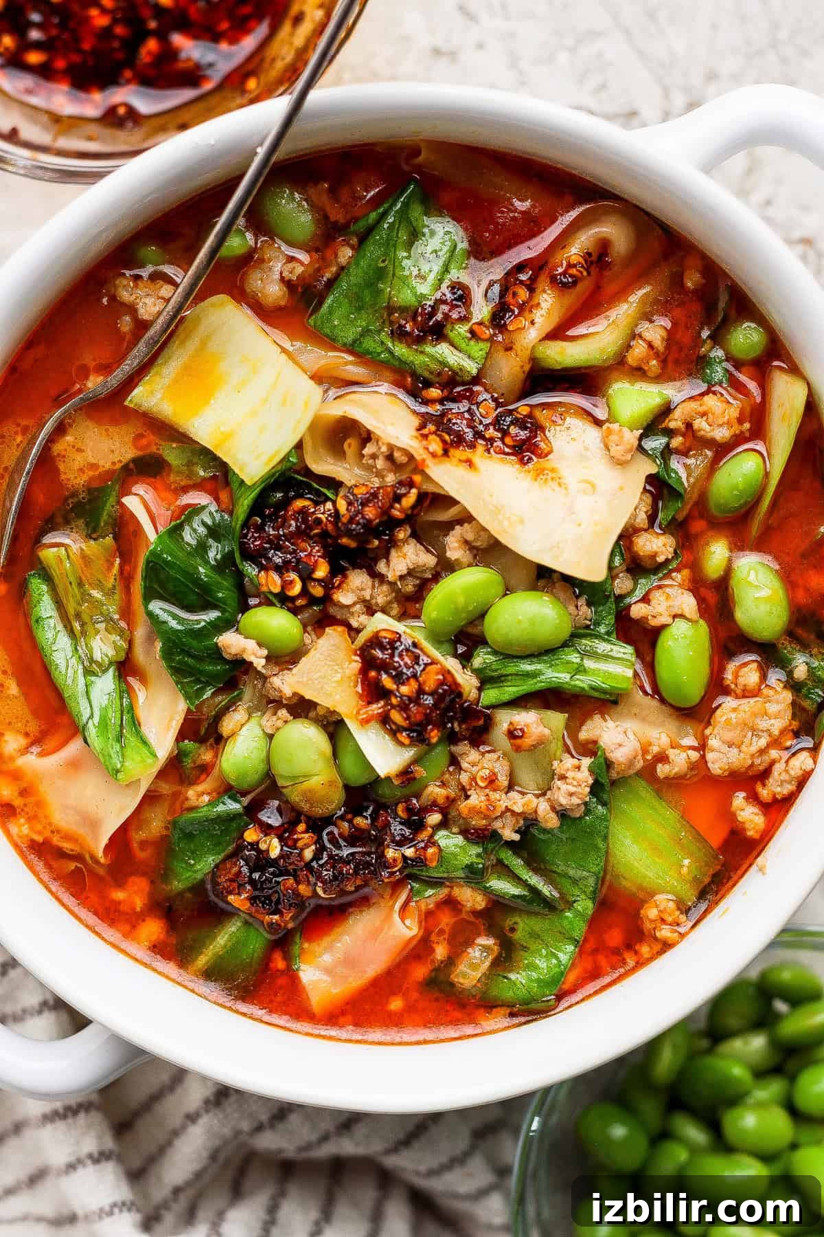 20-Minute Thai Coconut Dumpling Soup Recipe 2 A bowl of spicy noodle soup with ground meat, bok choy, edamame, chili oil, and wide noodles, served with a metal spoon.