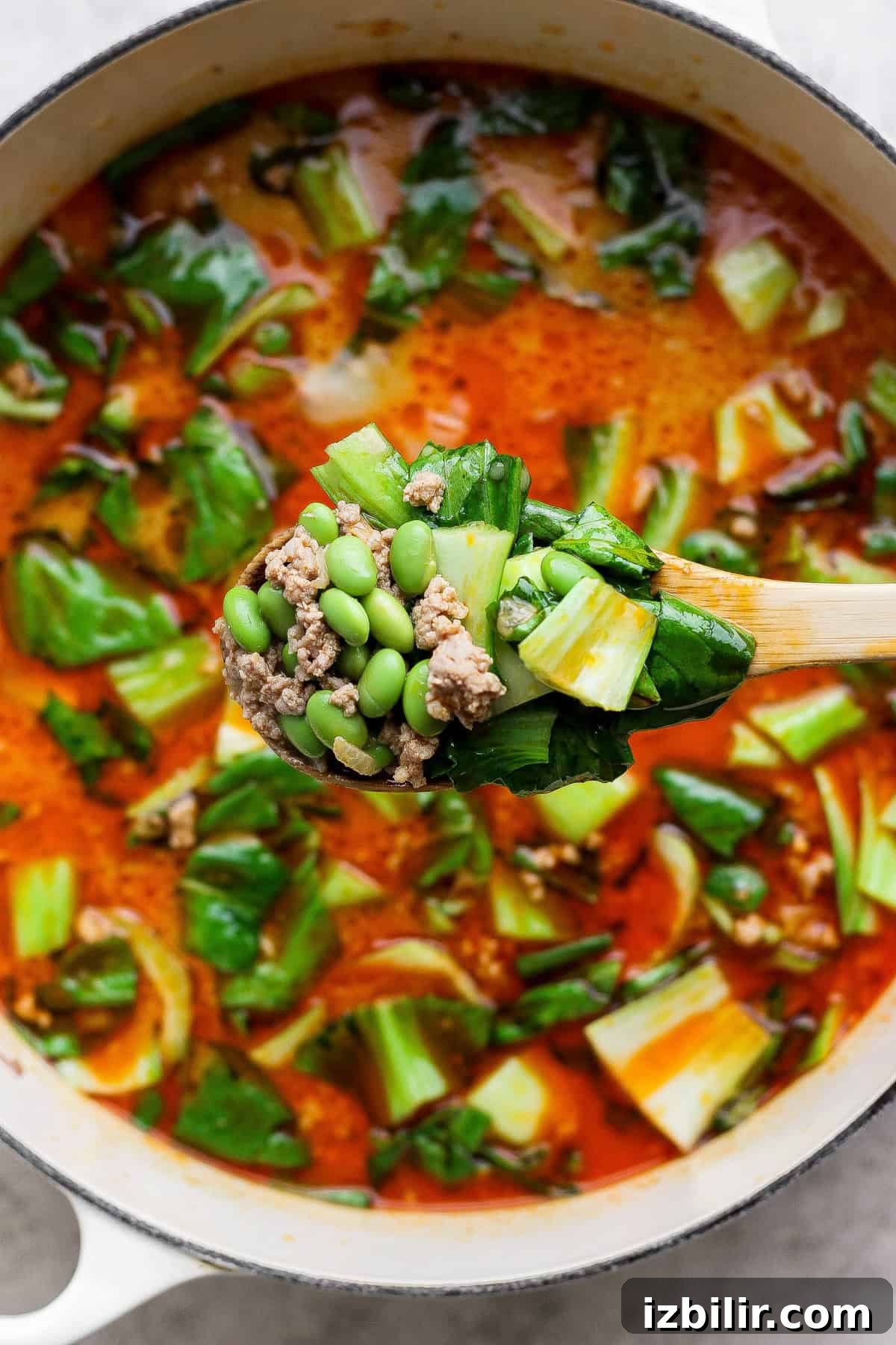 20-Minute Thai Coconut Dumpling Soup Recipe 4 A wooden spoon holds a mix of ground meat, edamame, and bok choy over a pot of red broth soup with leafy greens.