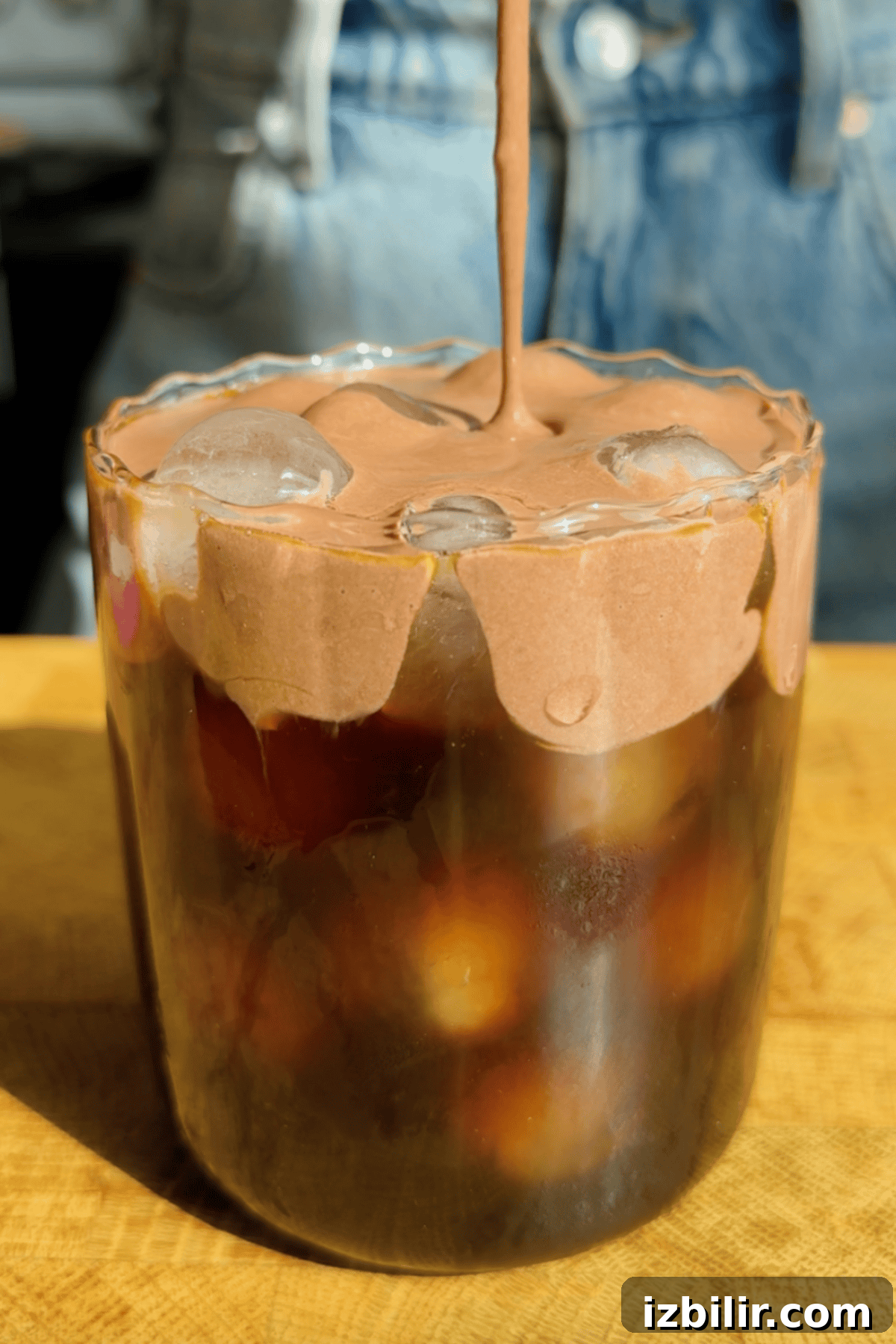 Starbucks Protein Cold Foam: Ingredients and How to Order 2 A glass of iced coffee topped with whipped chocolate cream, with someone pouring more cream on top. Denim clothing is visible in the background.