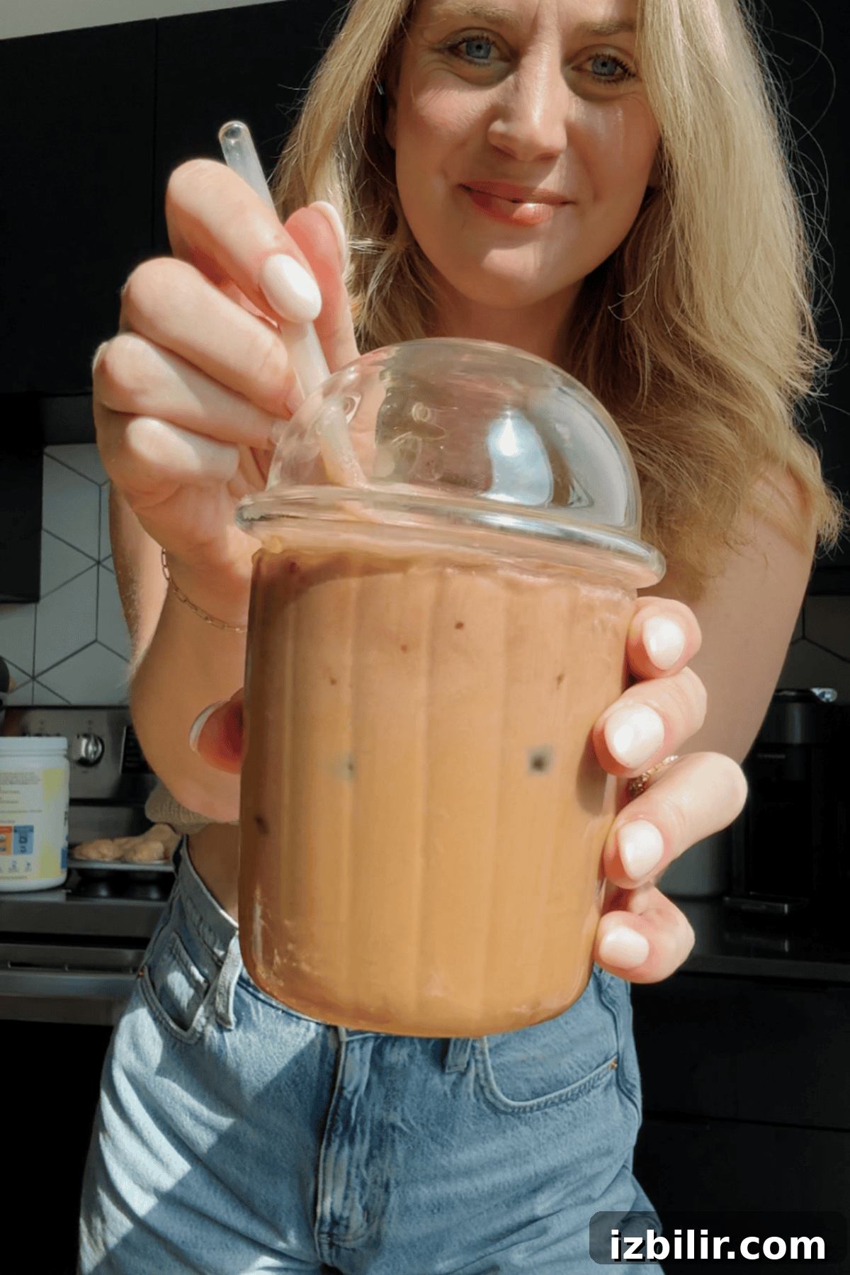 Starbucks Protein Cold Foam: Ingredients and How to Order 3 A woman holds a plastic cup of iced coffee with a dome lid and straw, topped with protein cold foam, standing in a kitchen.