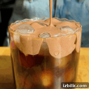 Starbucks Protein Cold Foam: Ingredients and How to Order 4 A glass of iced coffee topped with whipped chocolate foam, with the foam being poured over the ice. Denim fabric is visible in the background.