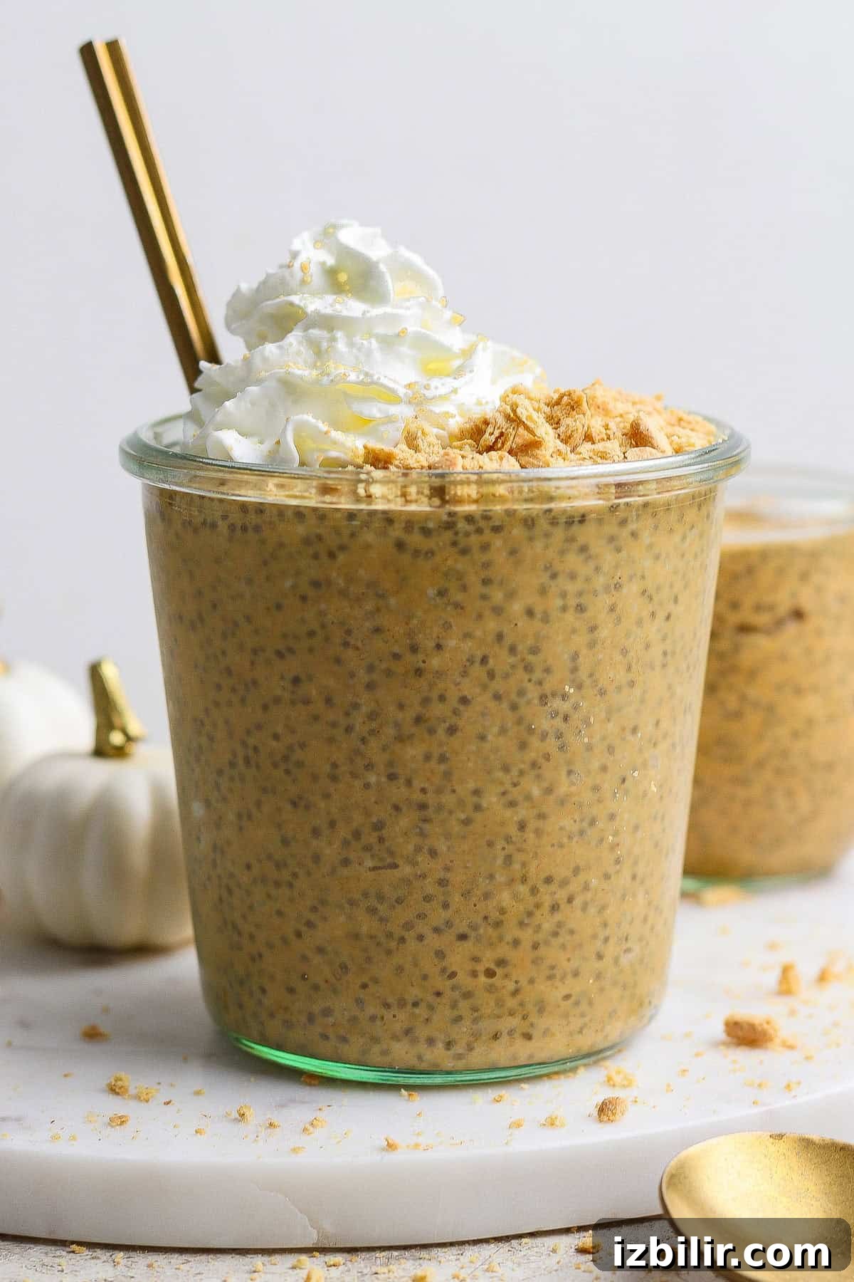 High-Protein Pumpkin Chia Pudding Recipe 5 A glass filled with pumpkin chia pudding, topped with whipped cream and crushed crumbs, with a gold spoon and straw, on a white surface.