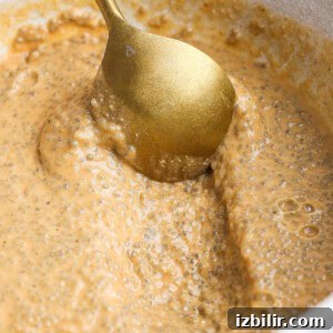 High-Protein Pumpkin Chia Pudding Recipe 8 A gold spoon stirs a thick, light brown chia pudding mixture in a white bowl.