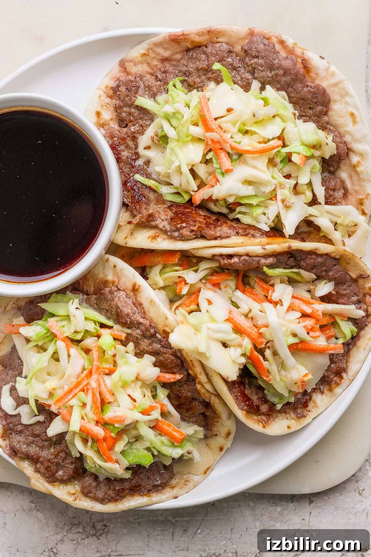 Three tacos filled with refried beans, shredded cabbage, and carrots are served on a white plate with a small bowl of dark dipping sauce.