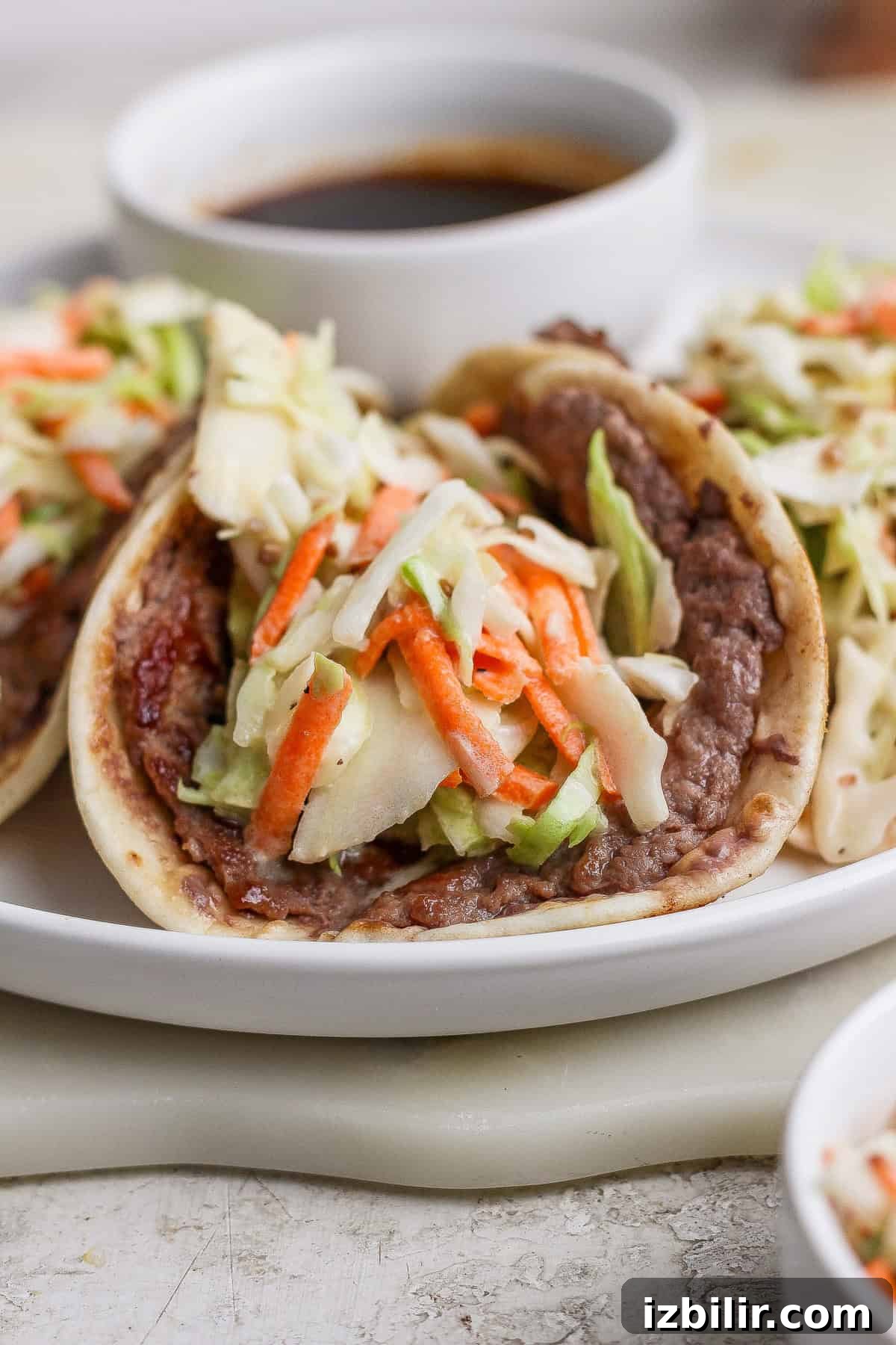 A soft taco filled with refried beans and topped with shredded cabbage and carrots is served on a white plate with a small bowl of sauce in the background.