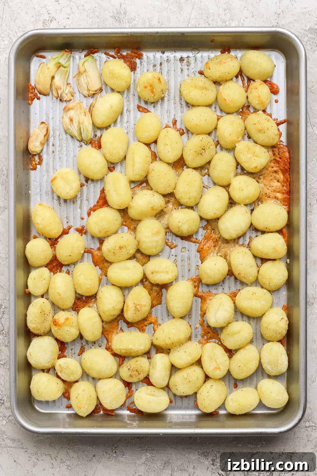 Garlic Parmesan Roasted Gnocchi and Kale Salad Recipe 3 A metal baking sheet with scattered cooked gnocchi, some clumps of melted cheese, and a few roasted garlic cloves on a textured surface.