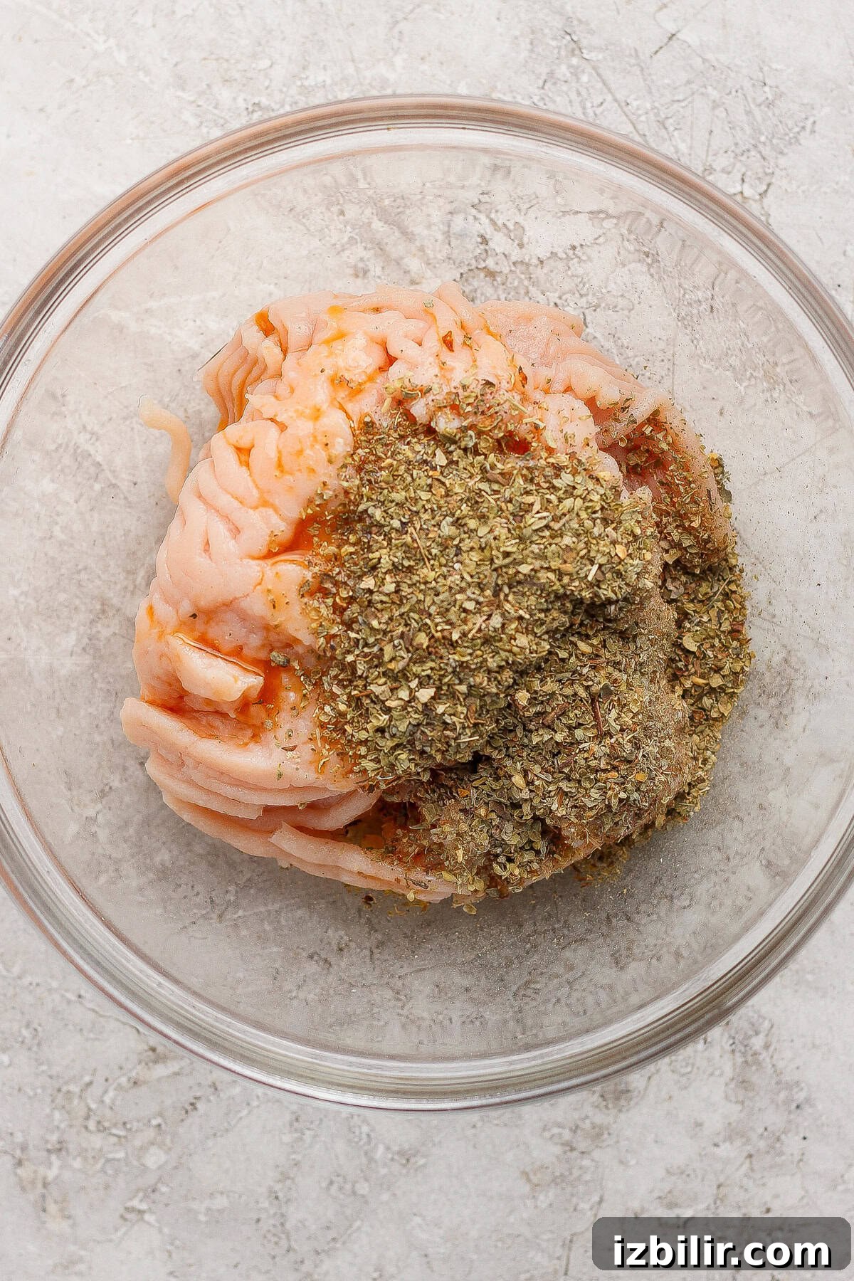 Raw ground meat topped with dried herbs and spices in a clear glass bowl on a light textured surface.