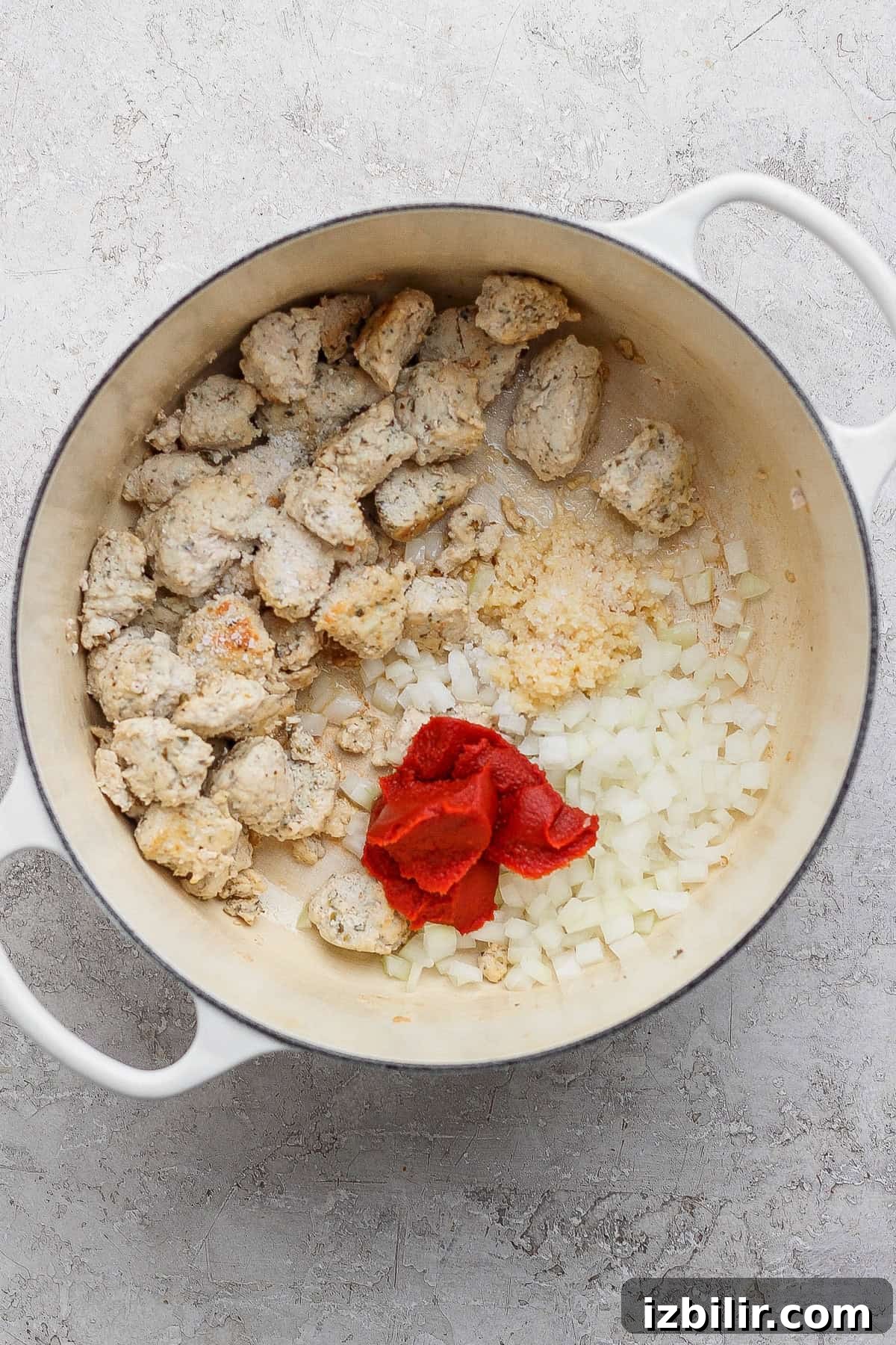 A white pot contains cooked chunks of sausage, chopped onions, minced garlic, and tomato paste on a light countertop.