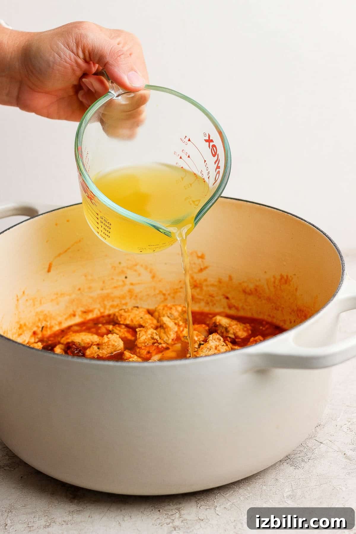 A hand pours chicken broth from a glass measuring cup into a white pot filled with cooked meat and sauce.