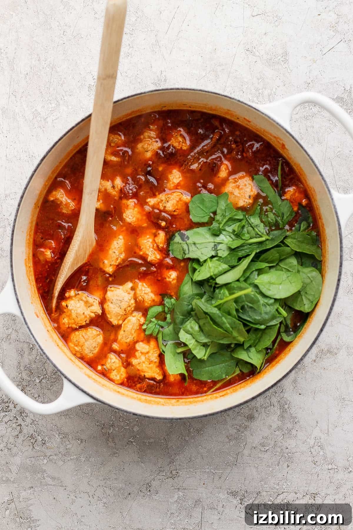 A white pot filled with tomato-based stew, meatballs, and fresh spinach, stirred with a wooden spoon on a light textured surface.