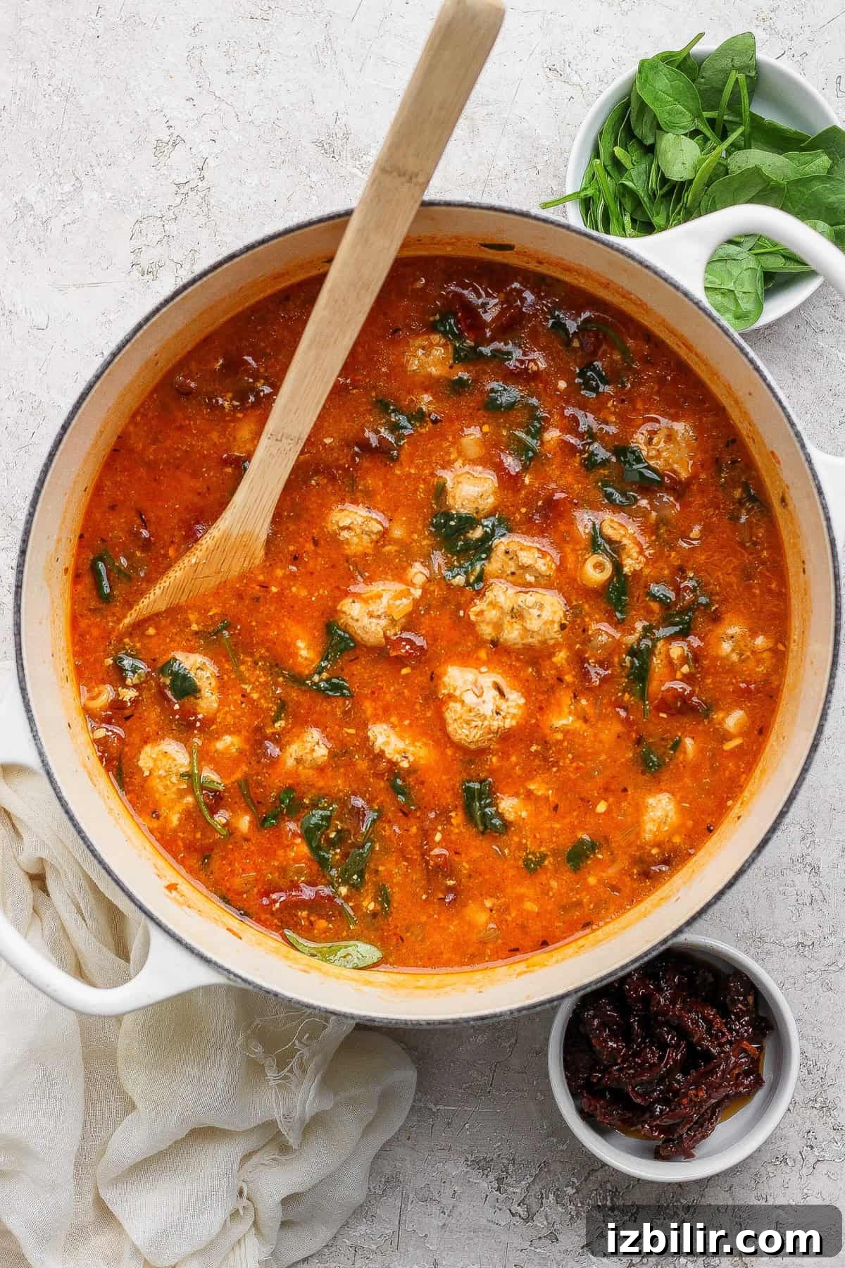 A pot of tomato-based soup with meatballs and spinach, a wooden spoon resting inside. Nearby are bowls of fresh spinach and sun-dried tomatoes.