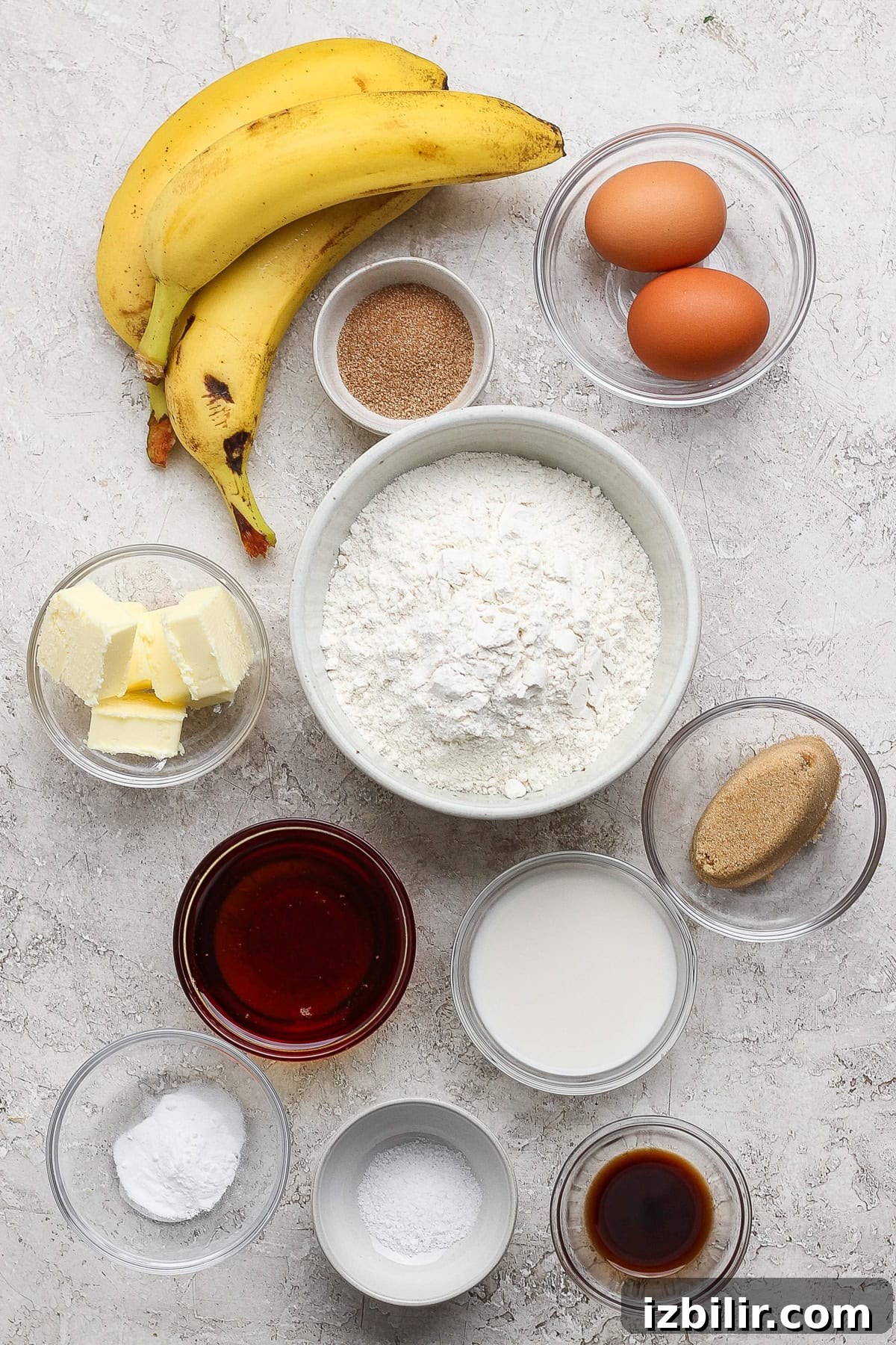 Banana bread recipe essentials: bananas, eggs, flour, sugar, butter, milk, brown sugar, baking powder, vanilla extract, salt, and cinnamon are beautifully arranged in bowls on a light surface.