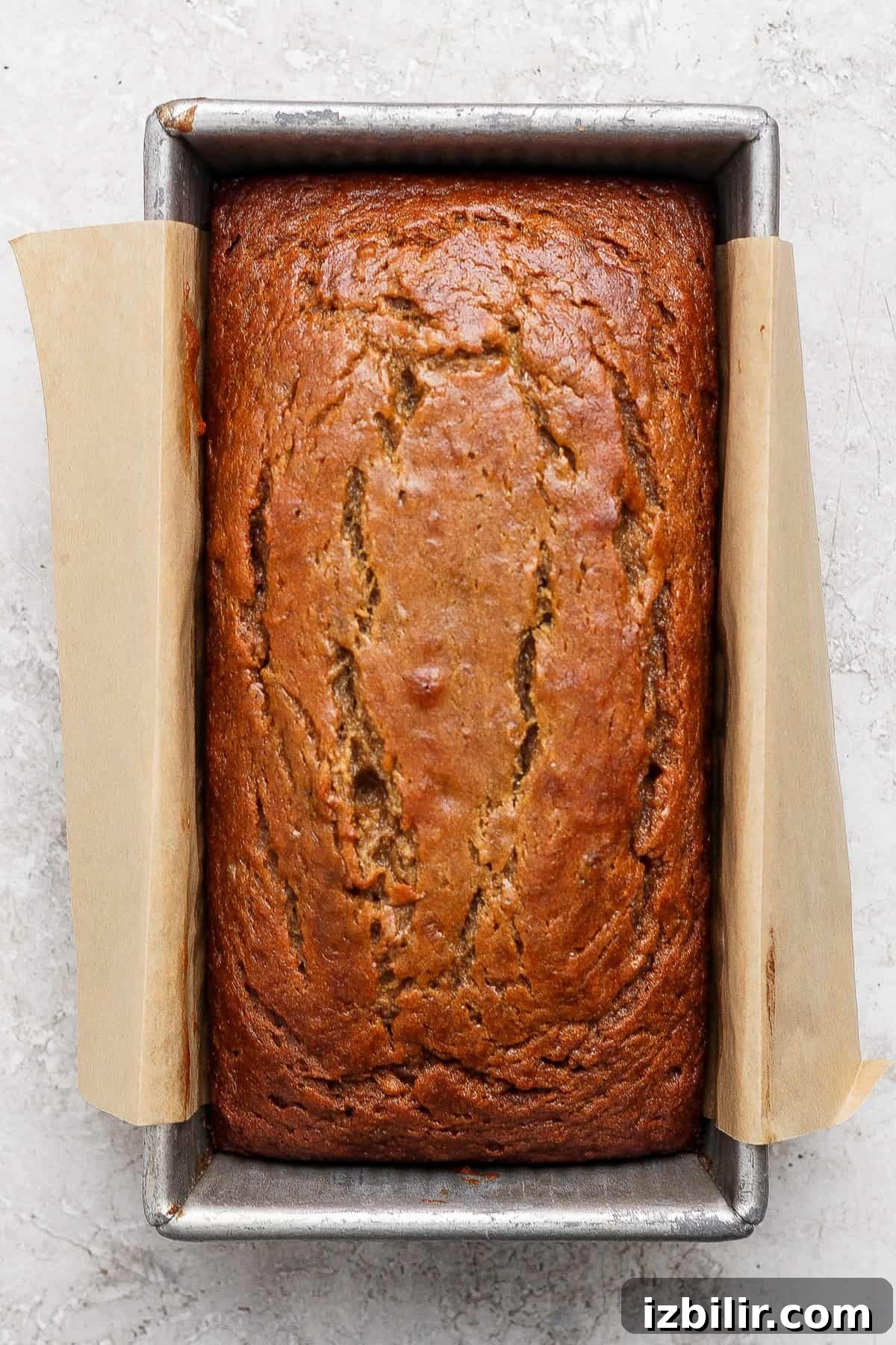 A loaf of banana bread from a classic banana bread recipe sits in a rectangular metal pan lined with parchment paper, shown from above on a light textured surface.