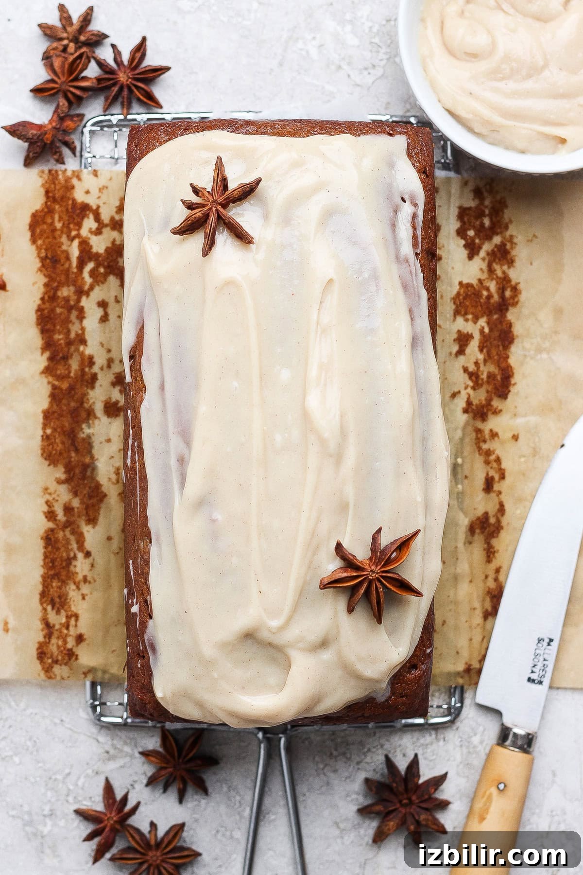 A delicious banana bread recipe loaf cake with light brown frosting, garnished with star anise, sits on parchment paper with a knife and a bowl of extra frosting nearby.