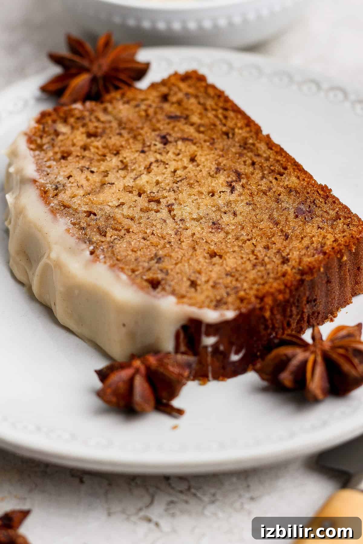 A slice of spiced banana bread loaf with white glaze on one edge, garnished with whole star anise, sits elegantly on a white plate—a delicious twist on the classic banana bread recipe.