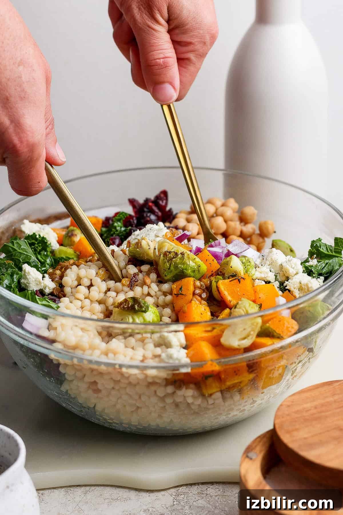 Autumn Couscous Salad Recipe with Seasonal Veggies 4 A person tossing a couscous salad recipe in a glass bowl with chickpeas, roasted vegetables, kale, red onion, cranberries, and cheese using two gold utensils.