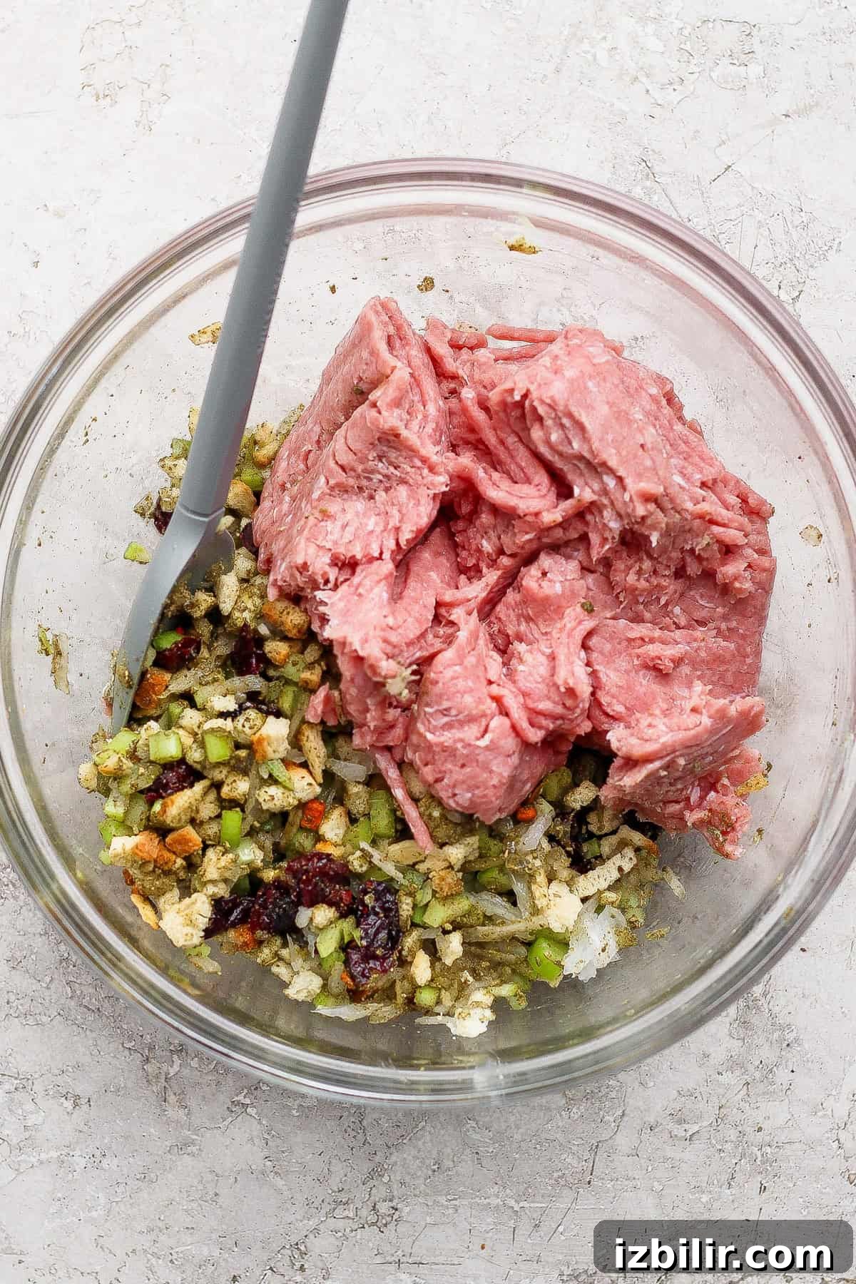 Thanksgiving Meatballs Recipe with Raspberry Sauce 3 A glass bowl containing raw ground meat, diced vegetables, bread cubes, and a gray mixing spoon on a light countertop.