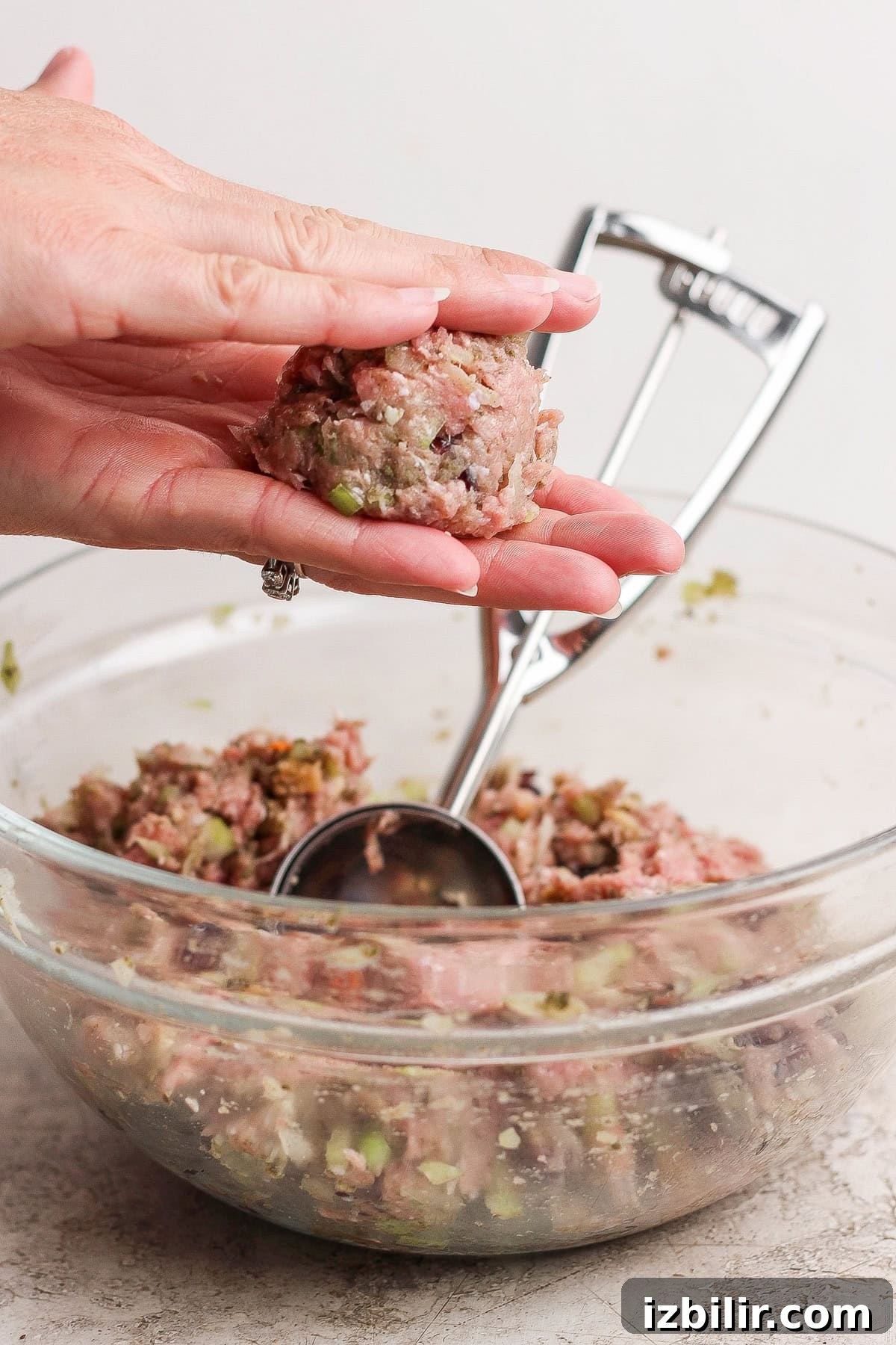 Thanksgiving Meatballs Recipe with Raspberry Sauce 4 A hand shapes a portion of raw meat mixture into a ball over a glass bowl filled with more mixture; a metal scoop rests in the bowl.