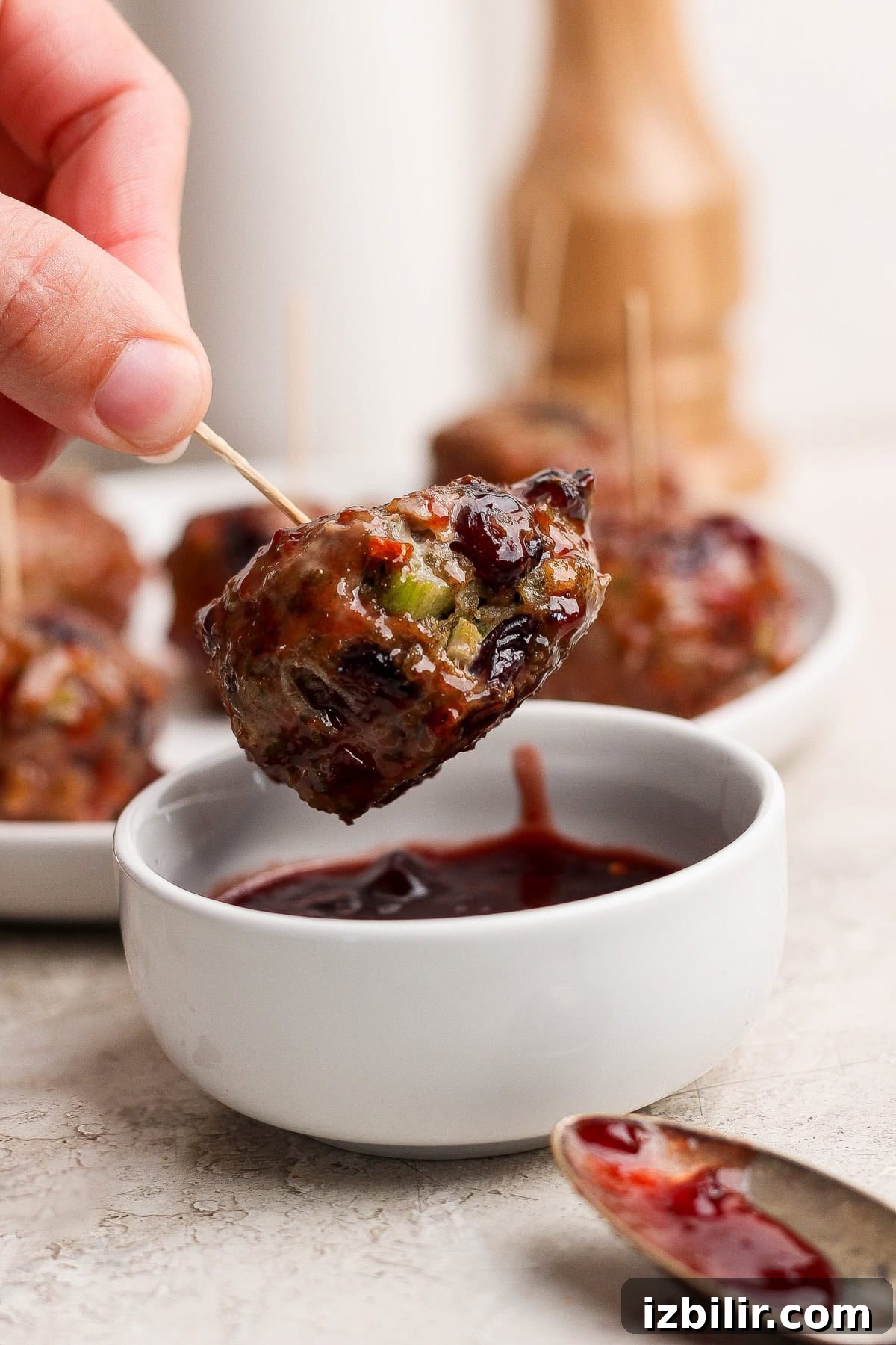 Thanksgiving Meatballs Recipe with Raspberry Sauce 7 A hand holds a meatball on a toothpick, dipping it into a small bowl of dark sauce, with more meatballs and a spoon nearby.