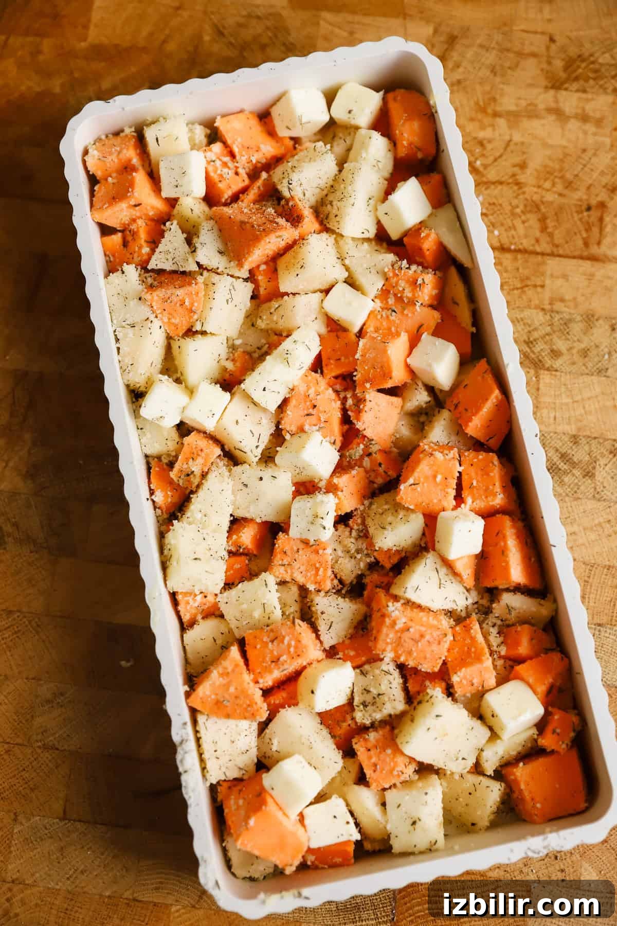 Sheet-Pan Thanksgiving Dinner for 4: Complete One-Tray Menu 4 A rectangular baking dish filled with cubed sweet potatoes and white potatoes, topped with small cubes of butter and sprinkled with seasoning, on a wooden surface.