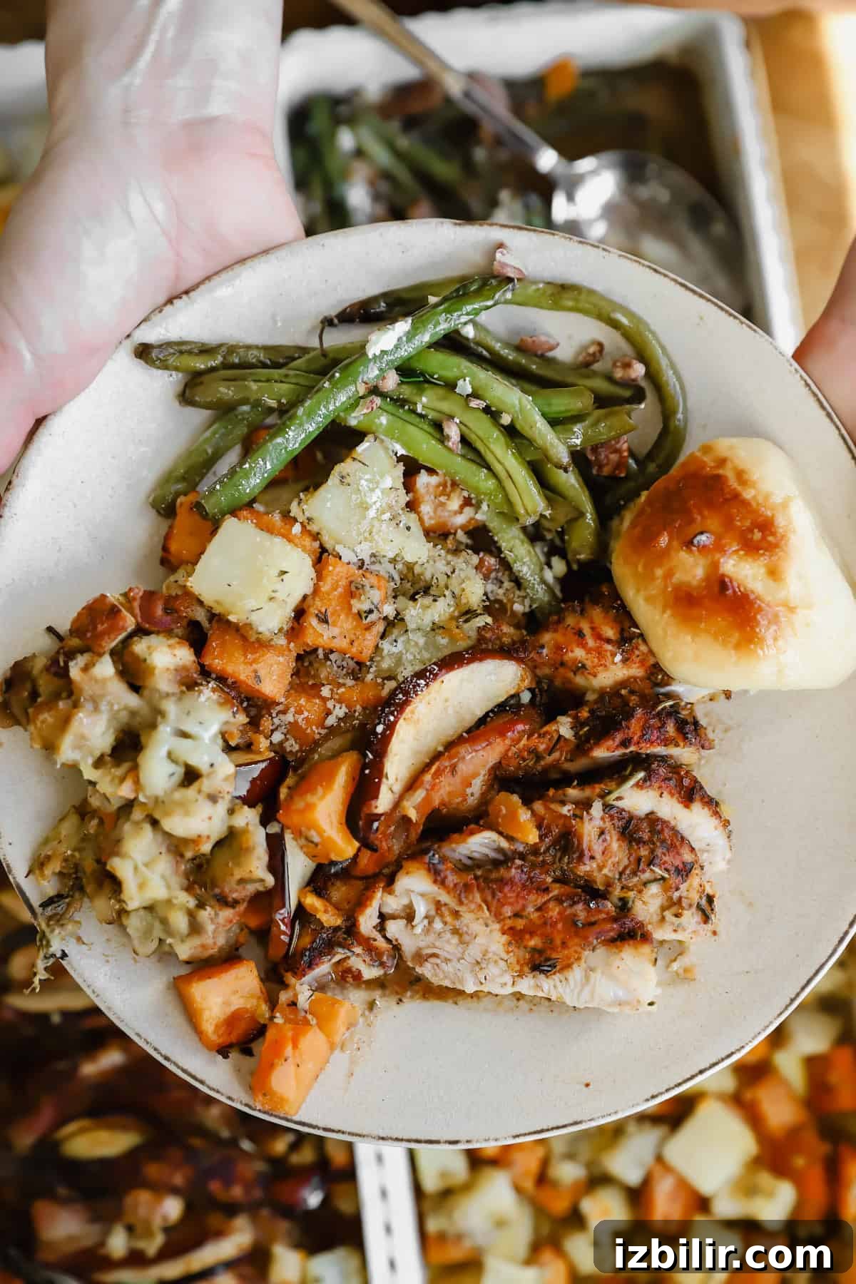 Sheet-Pan Thanksgiving Dinner for 4: Complete One-Tray Menu 8 A plate with roasted turkey, green beans, stuffing, roasted vegetables, and a dinner roll, held over a table with more food trays.