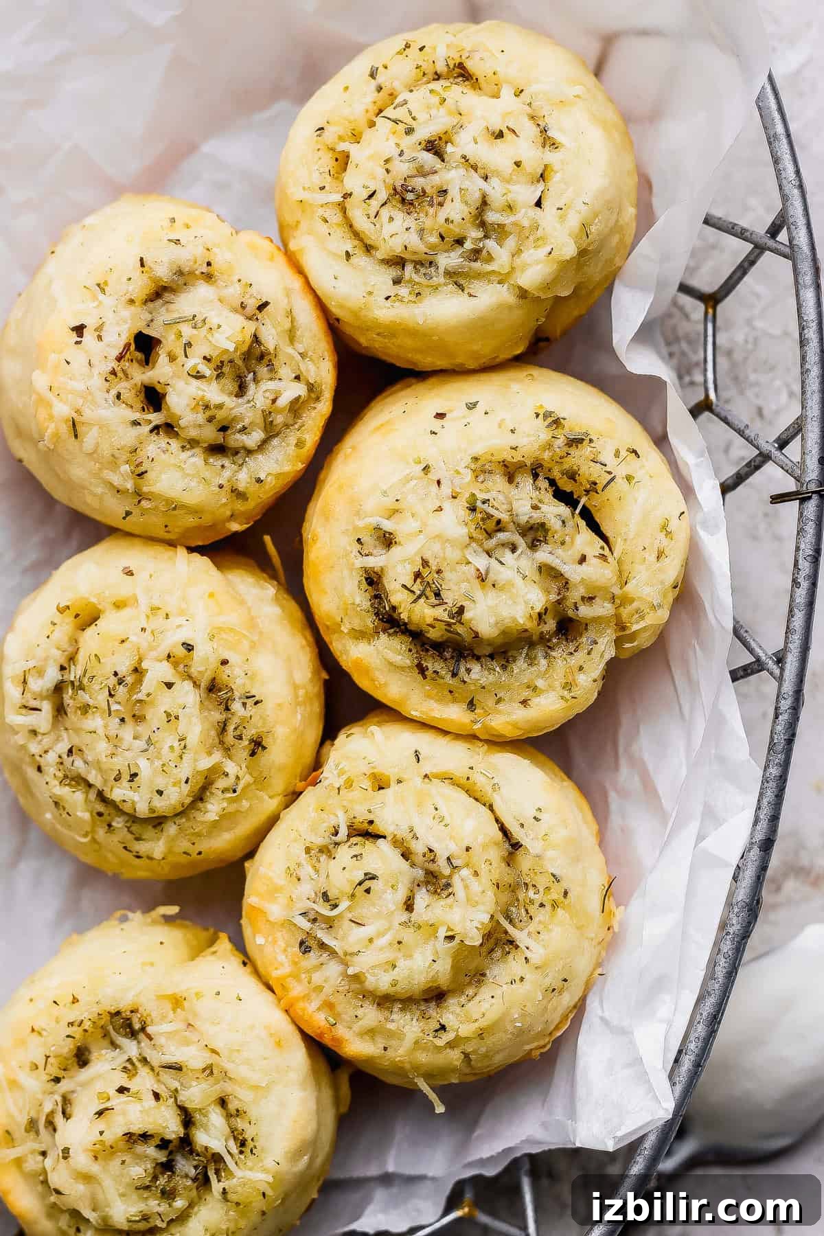 Protein-Packed Garlic Parmesan Cottage Cheese Dinner Rolls 2 Six golden-brown, spiral-shaped bread rolls topped with herbs and shredded cheese, arranged on white parchment paper in a wire basket.