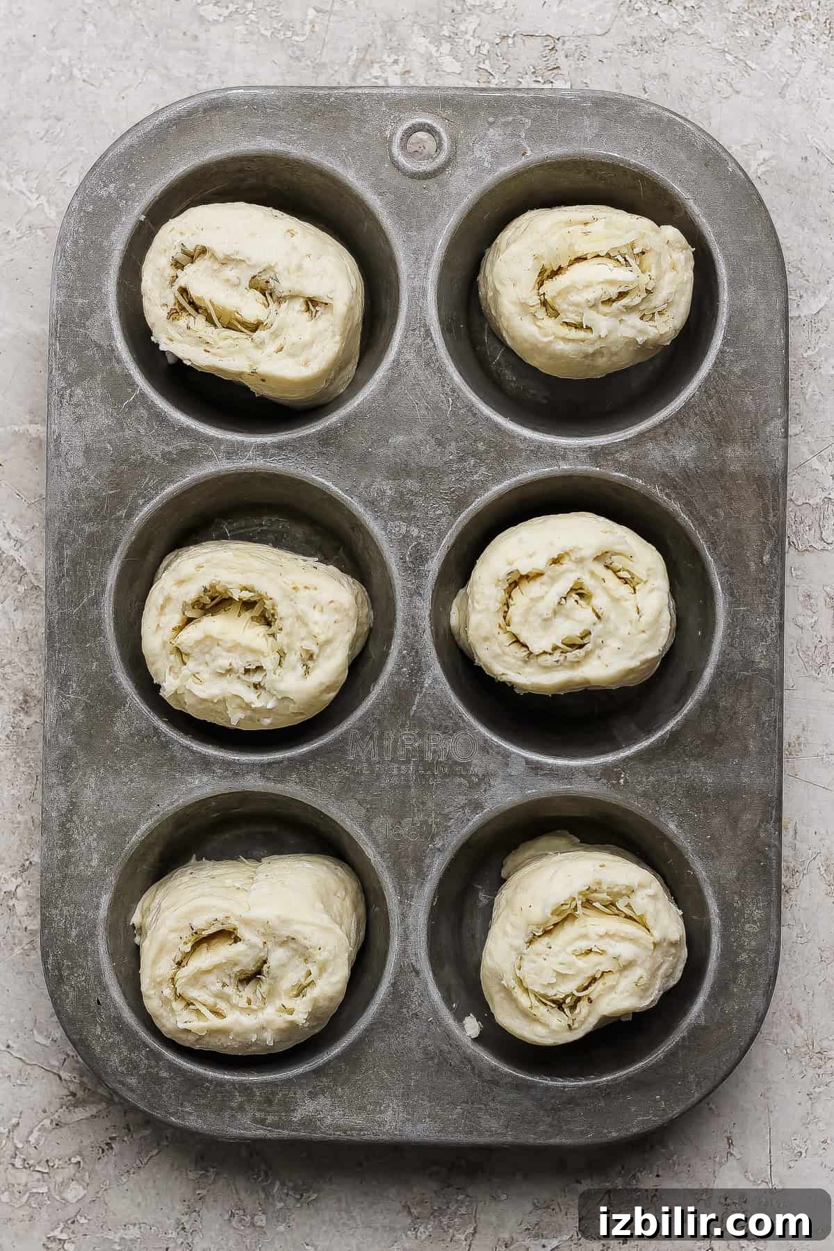 Protein-Packed Garlic Parmesan Cottage Cheese Dinner Rolls 5 Six unbaked rolled dough pieces placed in a metal muffin tin, ready for baking, on a textured light surface.