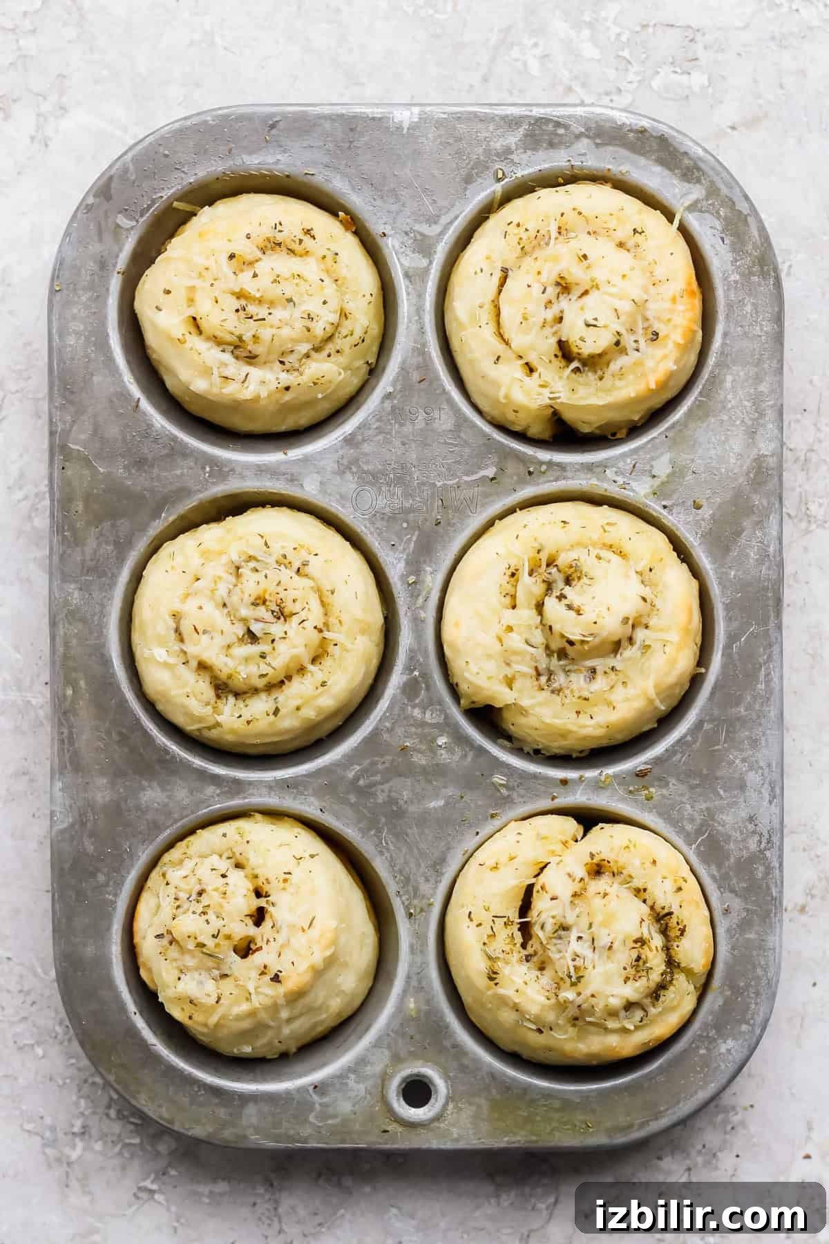 Protein-Packed Garlic Parmesan Cottage Cheese Dinner Rolls 6 A metal muffin tray holding six unbaked, spiral-shaped dough rolls topped with herbs and shredded cheese on a light textured surface.