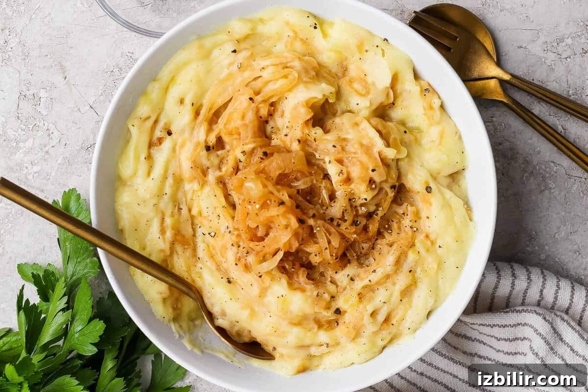 Creamy Mashed Potatoes with Caramelized Onions Recipe 2 A bowl of mashed potatoes topped with caramelized onions and black pepper, with a gold fork and spoon beside the bowl on a light textured surface.