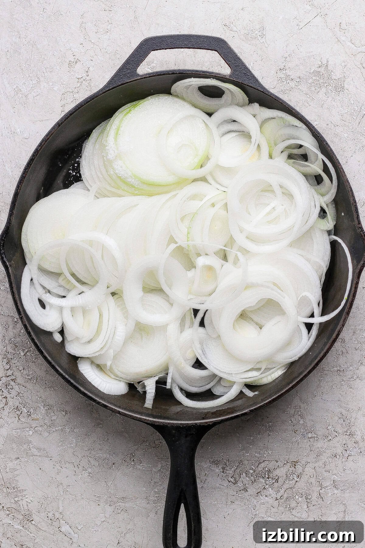 Creamy Mashed Potatoes with Caramelized Onions Recipe 3 A cast iron skillet filled with raw, sliced white onions, arranged in overlapping rings on a light textured surface.