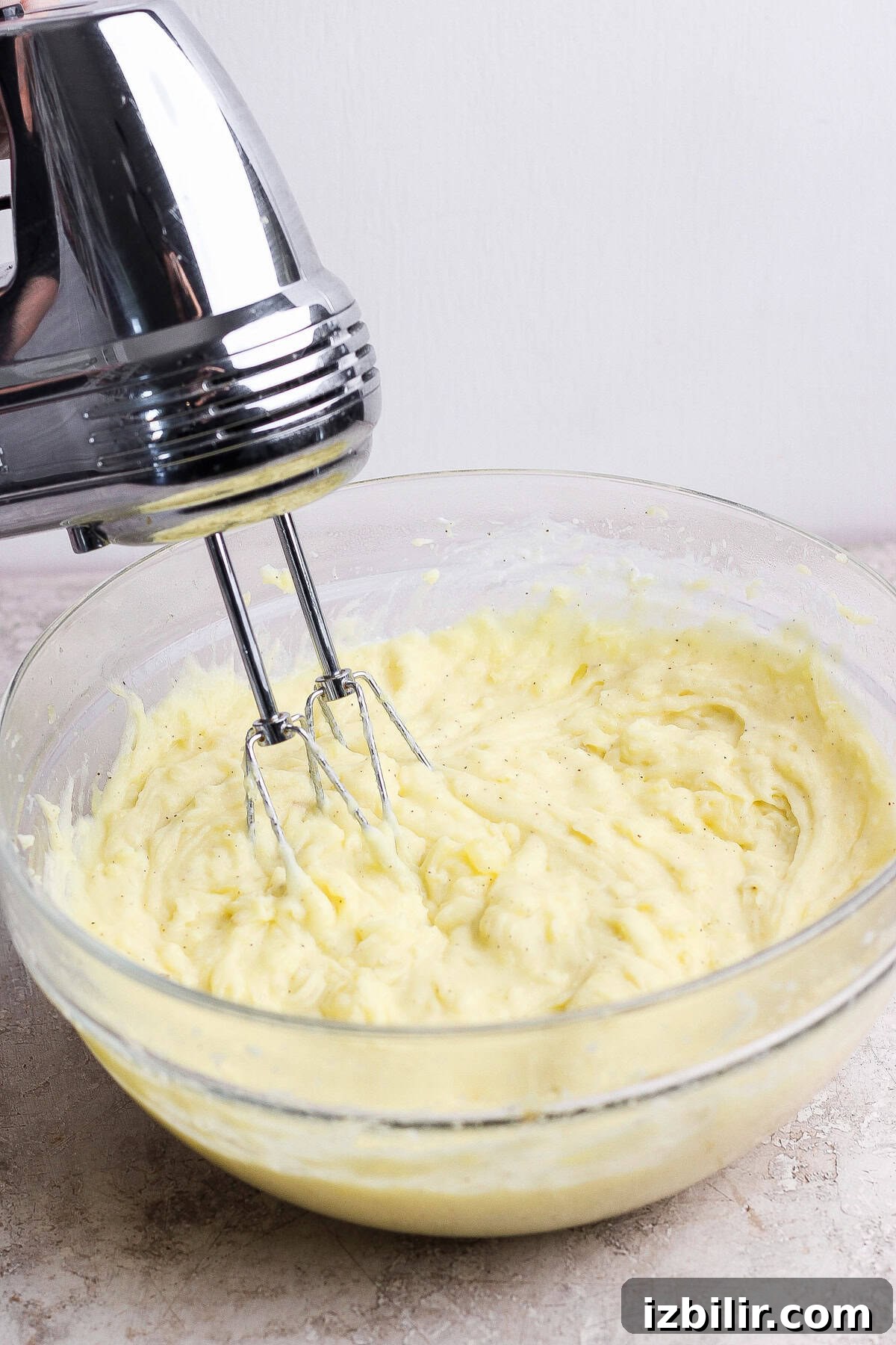 Creamy Mashed Potatoes with Caramelized Onions Recipe 6 A handheld electric mixer blending creamy batter in a clear glass bowl on a light counter.