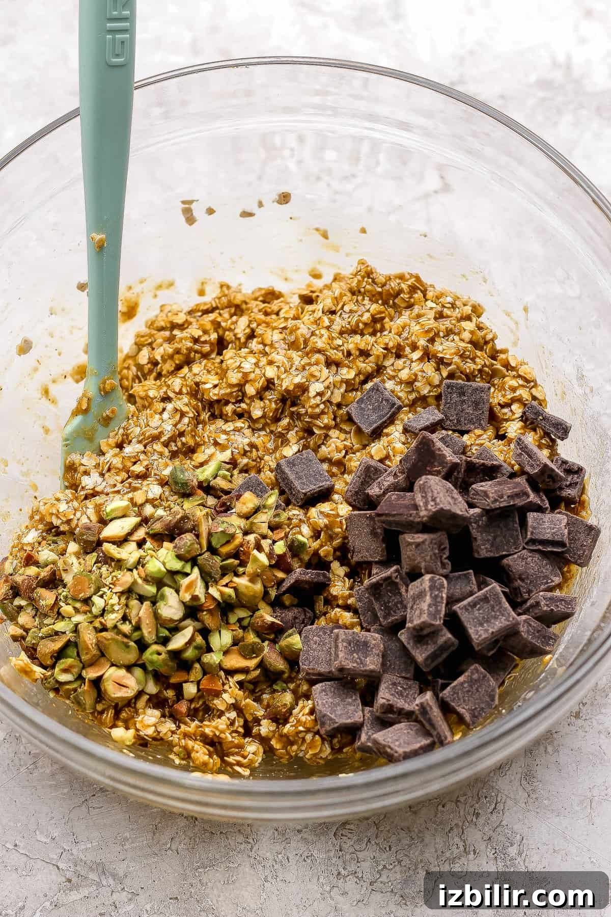 Chocolate Chunk Pistachio Cookies Recipe 3 A glass bowl containing oat mixture with chopped pistachios and dark chocolate chunks, and a green spatula resting inside.