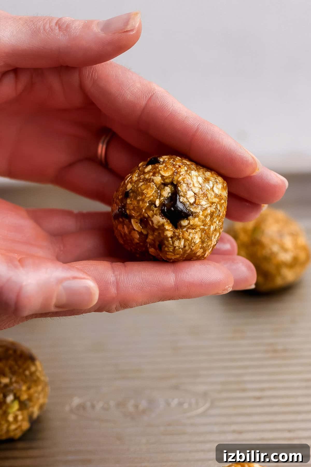 Chocolate Chunk Pistachio Cookies Recipe 4 A hand holding a round, oat-based energy bite with a visible chocolate chip, above a metal baking tray with more energy bites in the background.