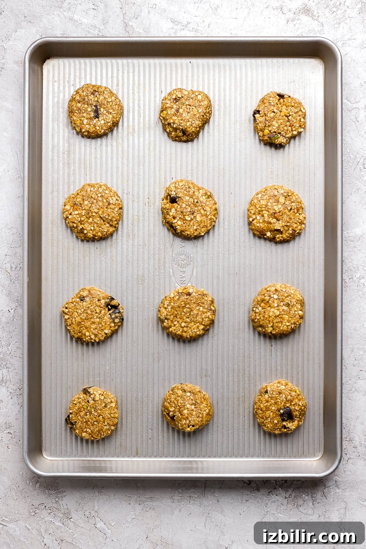 Chocolate Chunk Pistachio Cookies Recipe 5 Twelve unbaked oatmeal cookies with chocolate chips are evenly spaced on a metal baking sheet, ready to be baked.