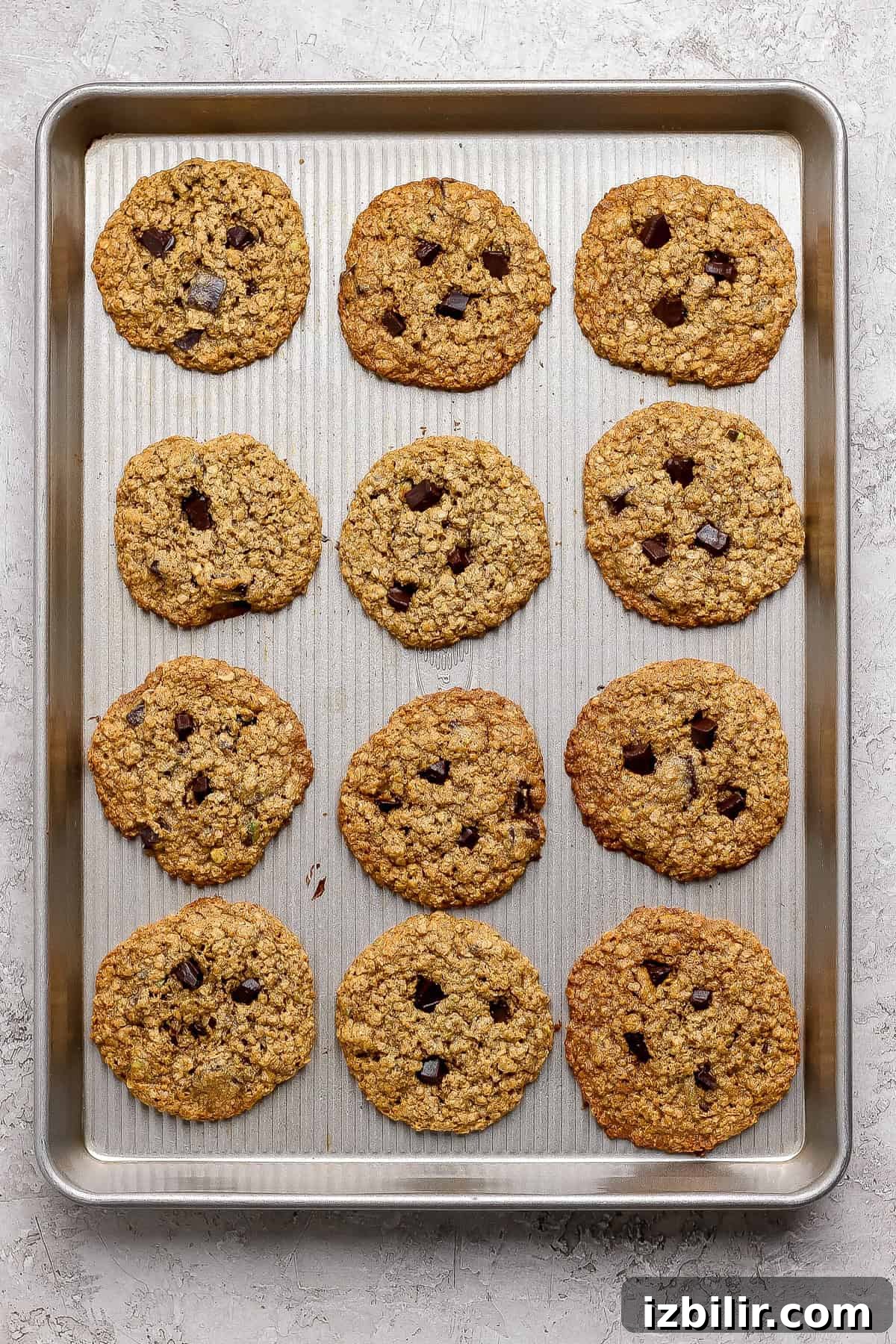 Chocolate Chunk Pistachio Cookies Recipe 6 A baking sheet holds twelve baked oatmeal chocolate chip cookies arranged in three rows of four on a light-colored surface.