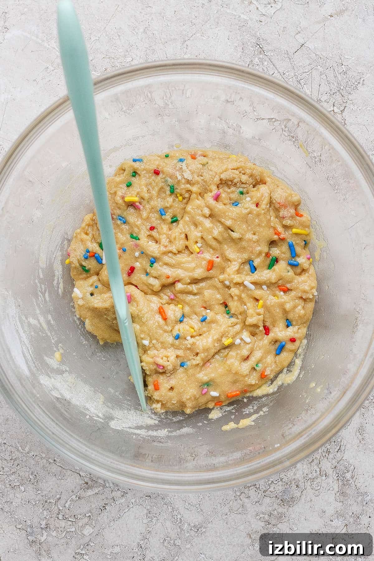 A glass bowl with raw cookie dough and colorful sprinkles and a light blue spatula.