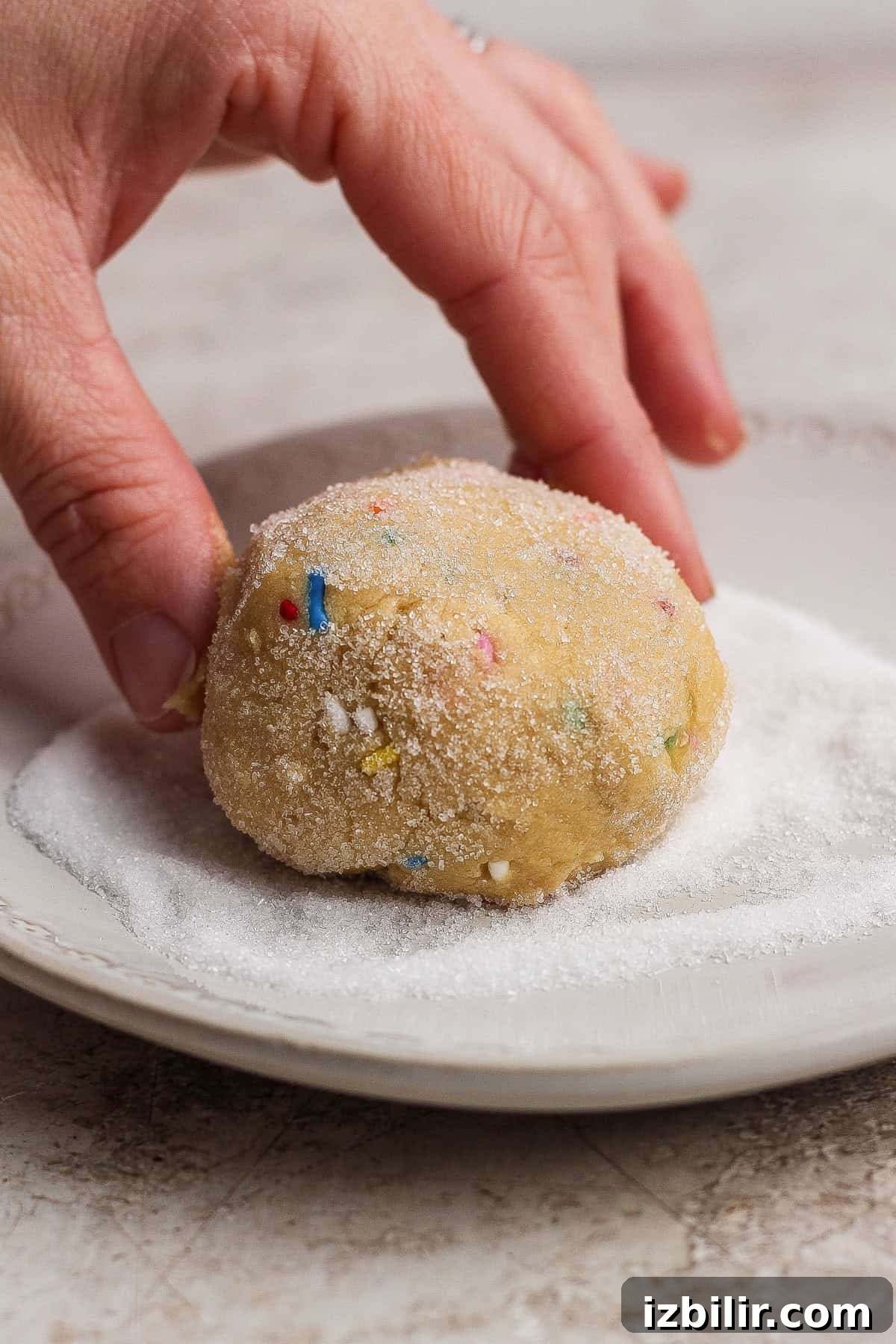 A hand rolling a ball of cookie dough with colorful sprinkles in a plate of sugar.