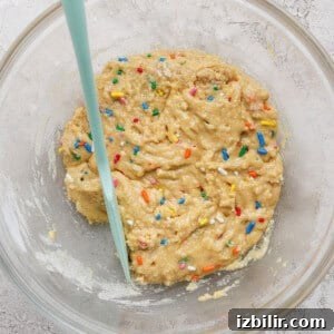 A glass bowl containing raw cookie dough with colorful sprinkles and a light blue spatula.