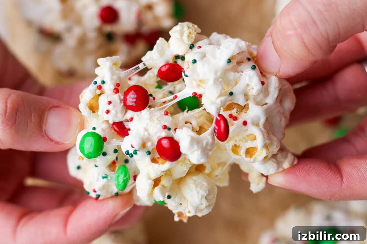 Close-up of hands holding a popcorn ball decorated with red and green candy-coated chocolates and festive sprinkles.