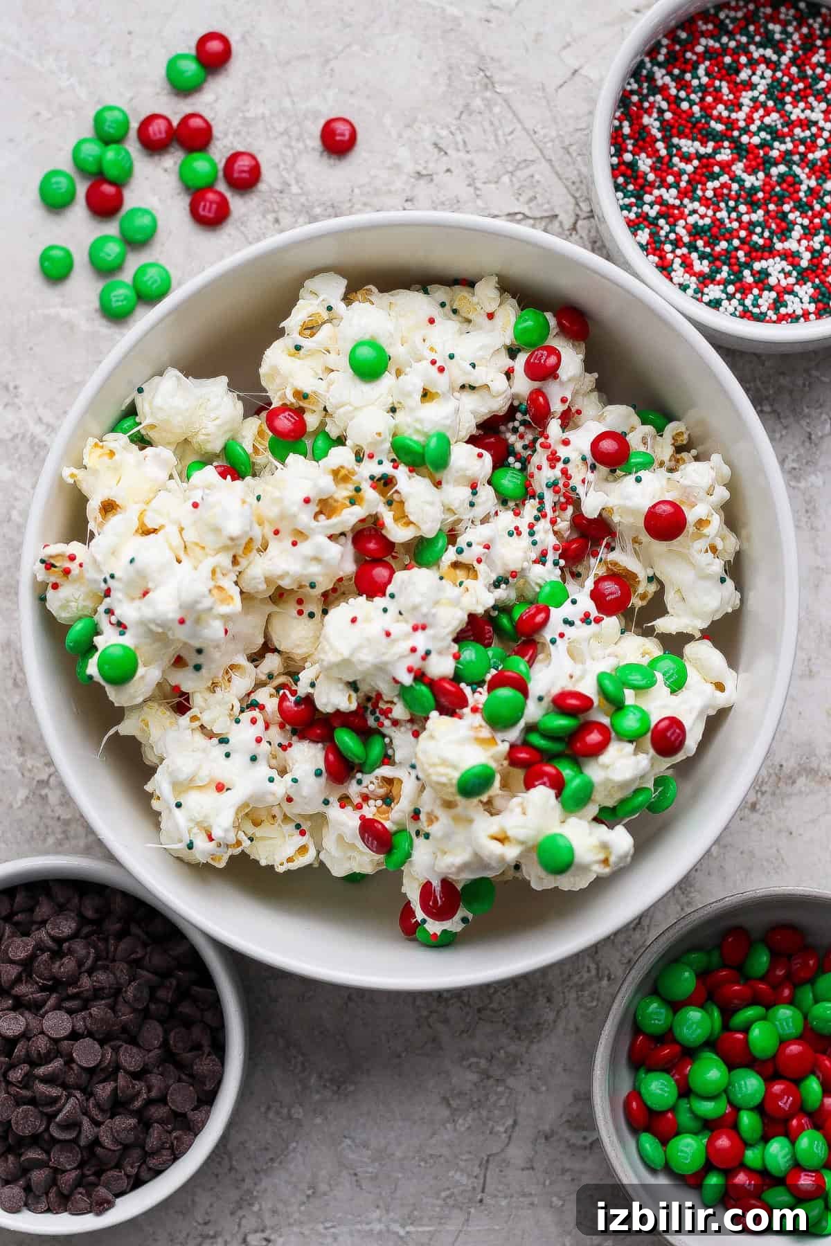 A bowl of popcorn topped with red and green candy-coated chocolates and sprinkles, surrounded by bowls of chocolate chips, more candies, and sprinkles on a light surface.