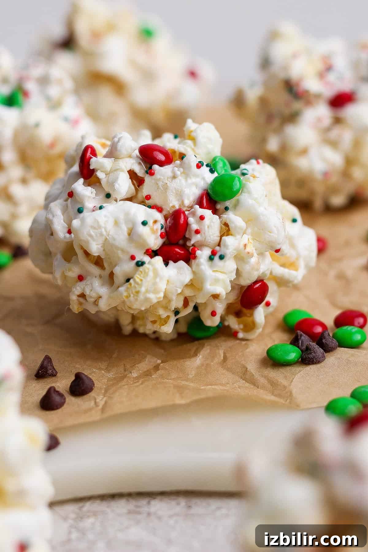 A close-up of a popcorn ball decorated with red and green candy-coated chocolates and multicolored sprinkles, placed on brown parchment paper.