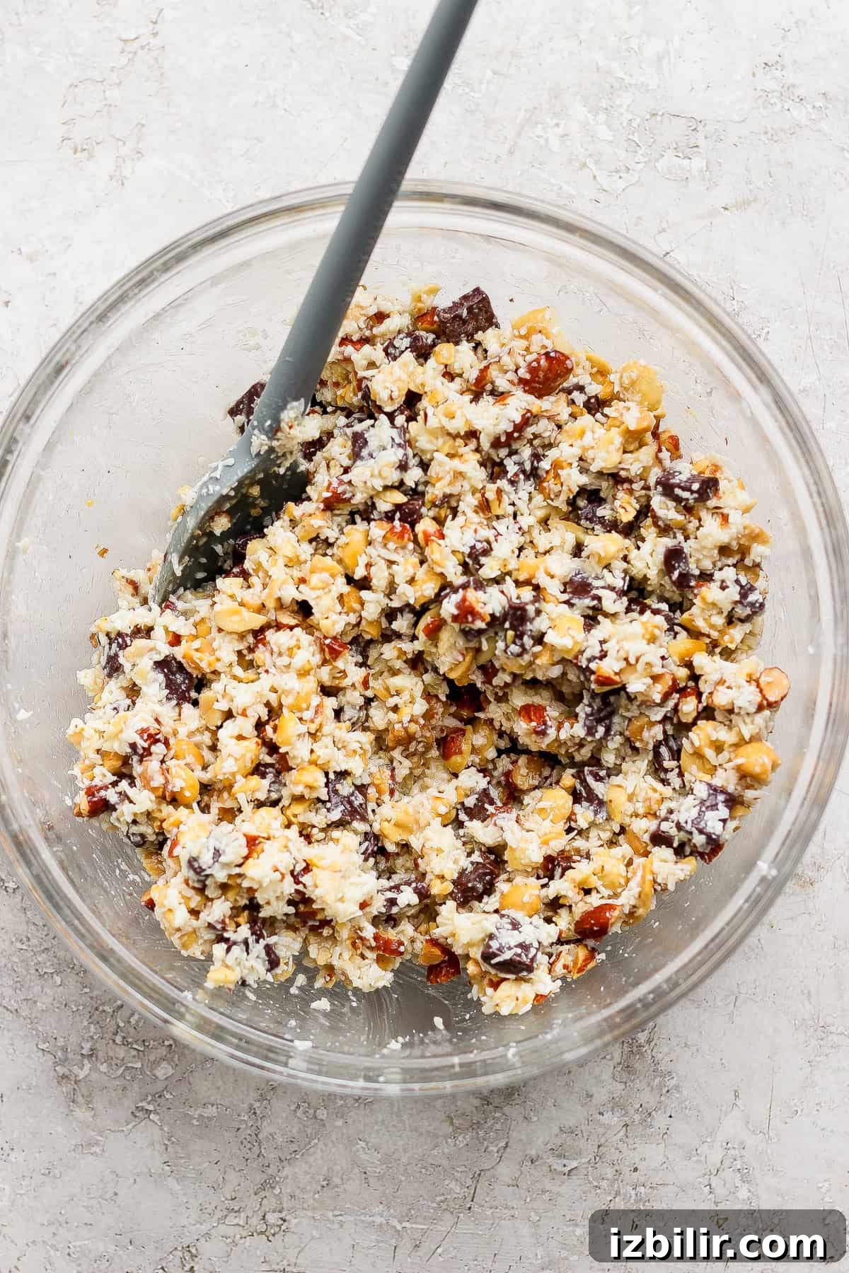 A glass bowl filled with a mixture of chopped nuts, chocolate chunks, and coconut flakes, with a gray spatula resting inside.