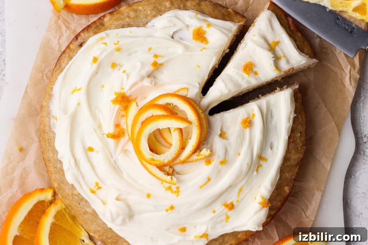 A round cake with white frosting and orange zest, topped with orange peel spirals, has one slice being removed; orange slices are placed beside the cake.