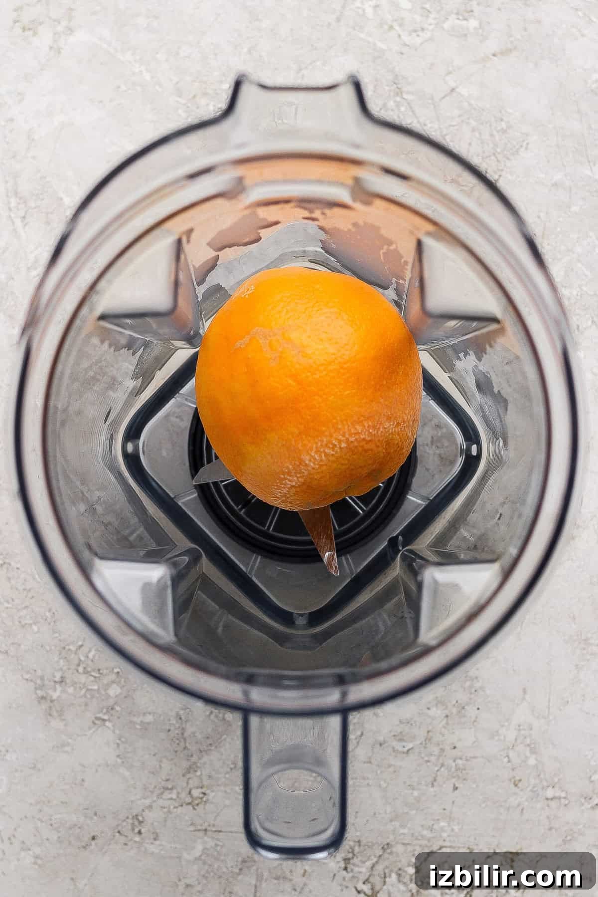 A whole orange with a small peel removed sits at the bottom of an empty blender viewed from above.