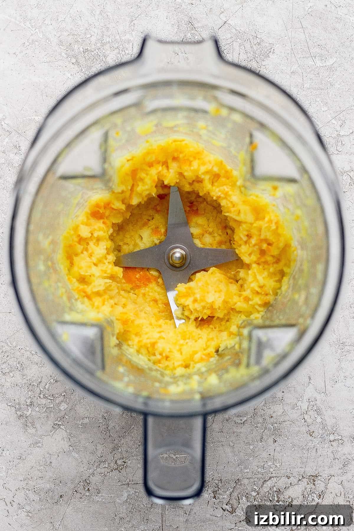 Top-down view of a blender with a yellow-orange mixture partially pureed, showing metal blades and textured sides, on a light gray surface.