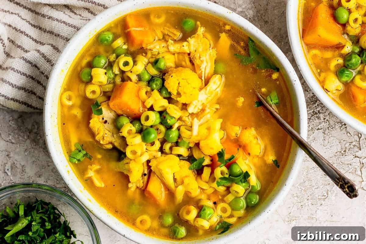 Comforting Soup to Boost Immunity 2 A bowl of yellow soup with small pasta, peas, cauliflower, and chunks of orange vegetables, garnished with chopped herbs, and a spoon inside the bowl.