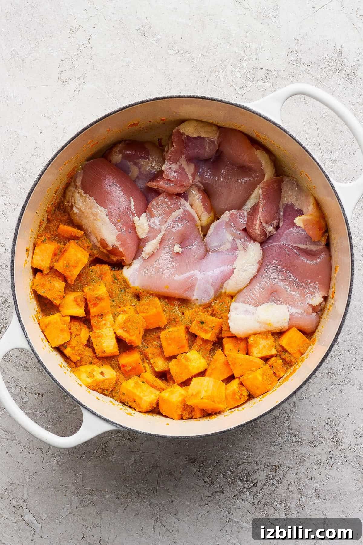 Comforting Soup to Boost Immunity 5 Raw chicken thighs and cubed sweet potatoes in a white Dutch oven on a light textured surface.